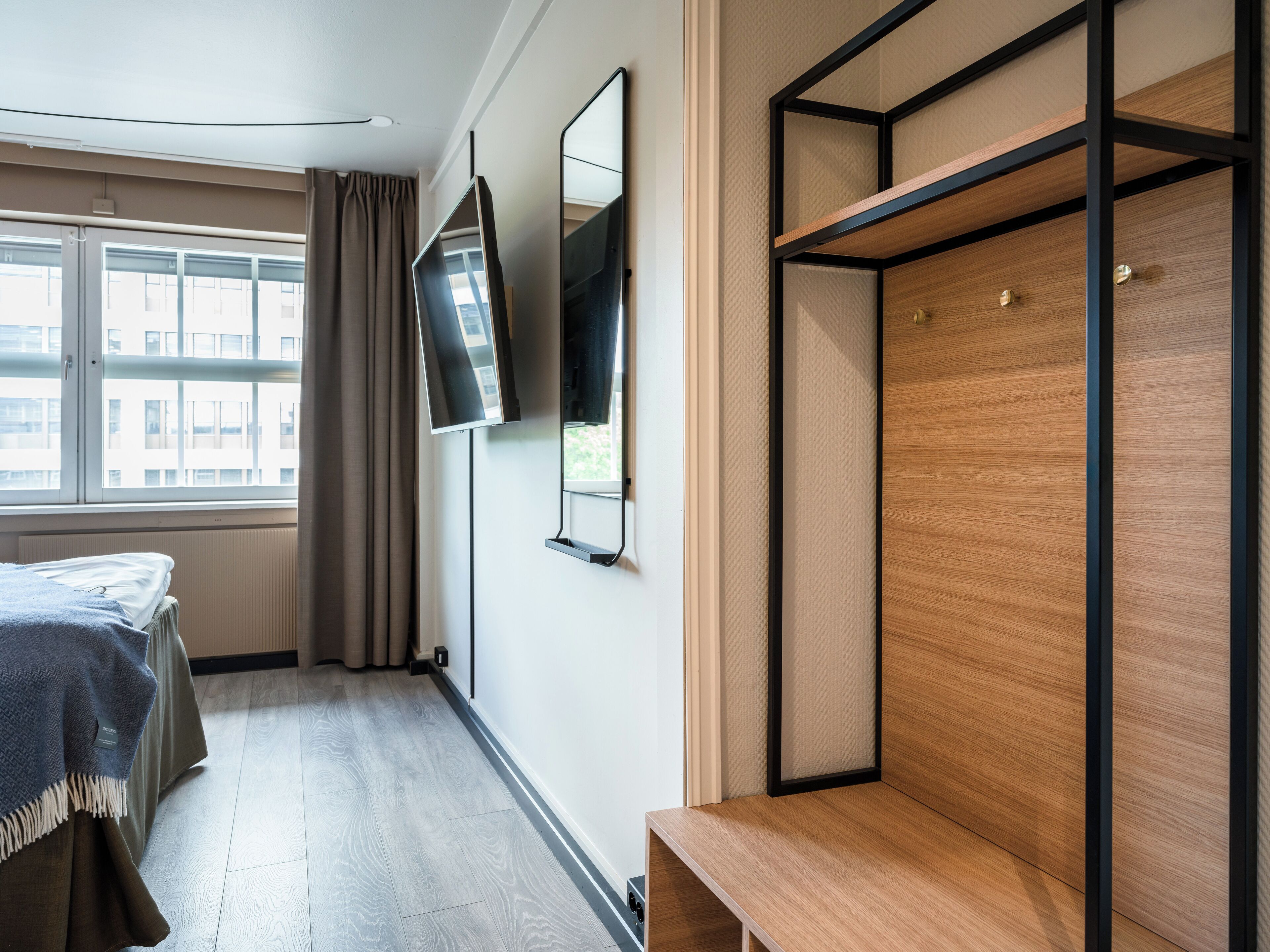 A hotel room with a bed, a TV mounted on the wall, a wooden bench, and a window with curtains.