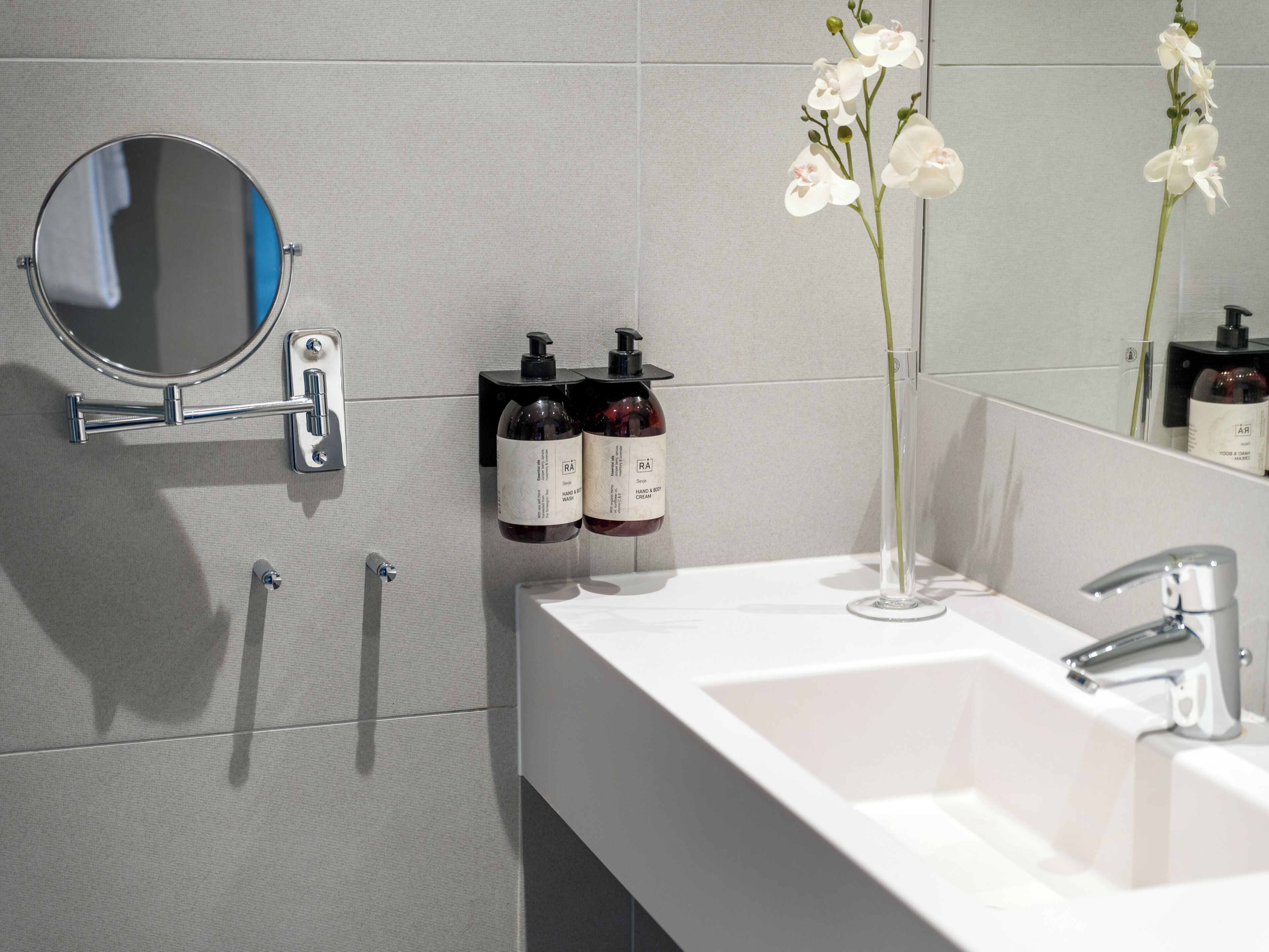 A bathroom with a white sink, a mirror, two hand and body wash bottles, and a vase with white flowers.