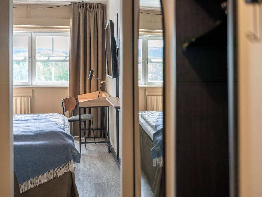 A room with two beds, a desk, and a chair. There is a window with a view of greenery.