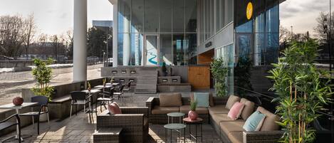 An outdoor seating area with wicker furniture and potted plants, a staircase leading to an entrance, and a modern building facade with large windows.