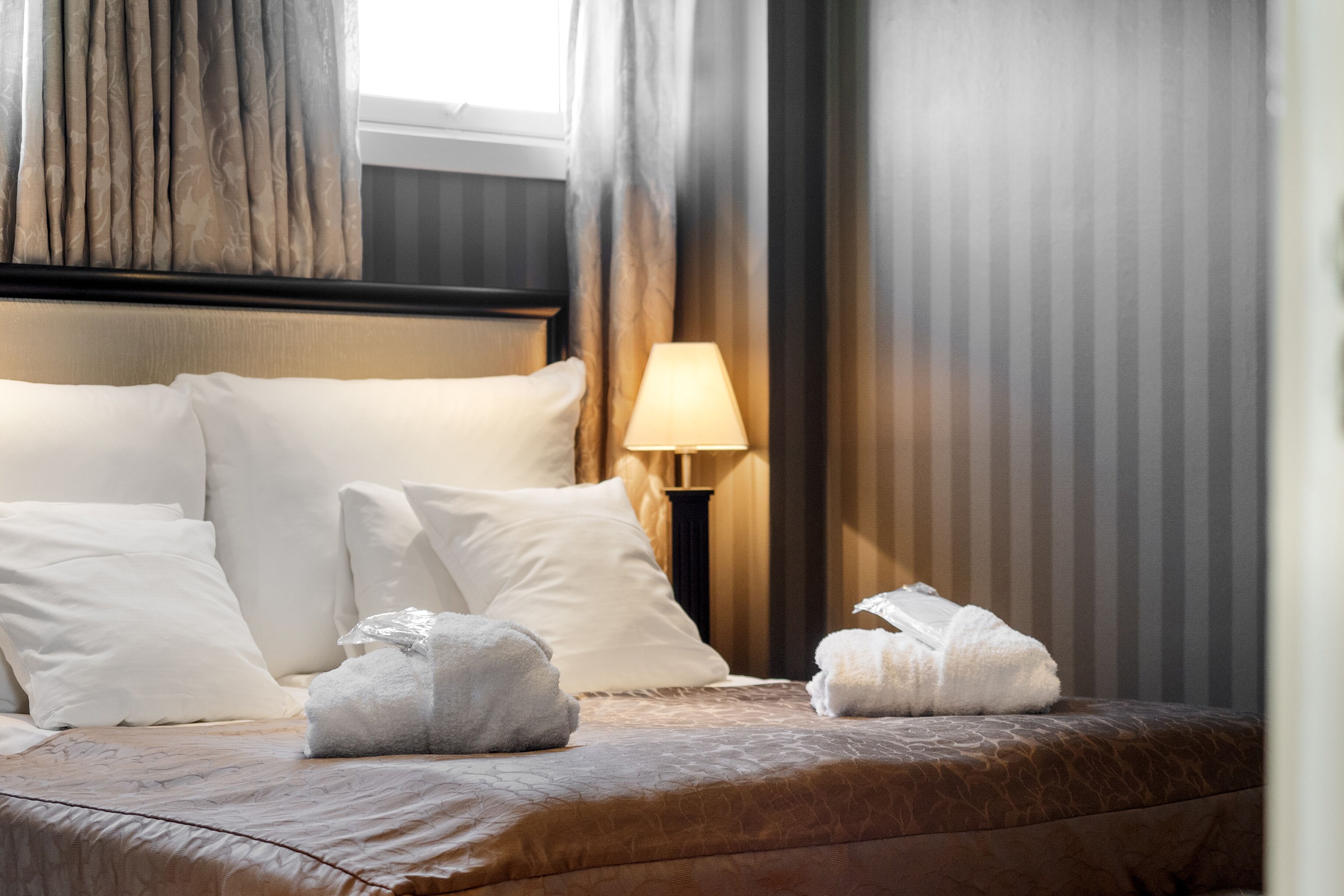 Una cama bien hecha con almohadas y toallas blancas, una lámpara de noche y una ventana con cortinas.