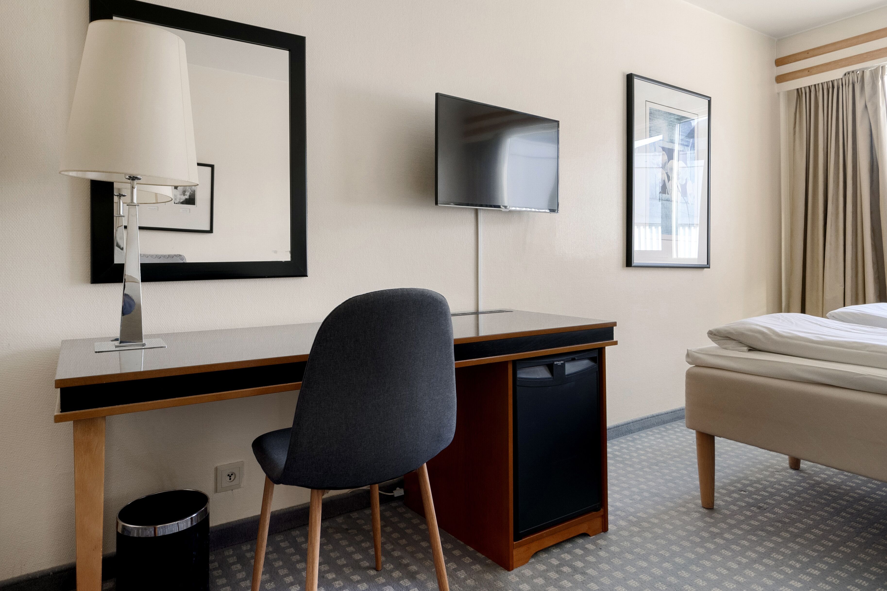 A hotel room with a desk, chair, TV, bed, and a small trash bin.