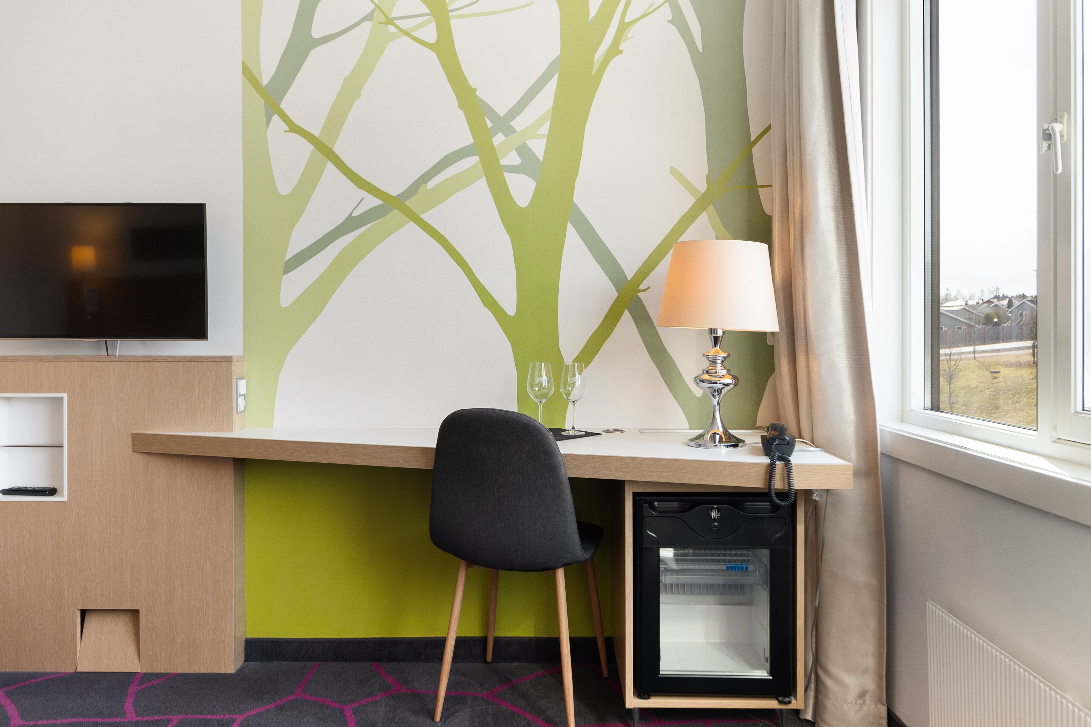 A hotel room with a desk, a chair, a TV, a lamp, a mini fridge, and a window with curtains.