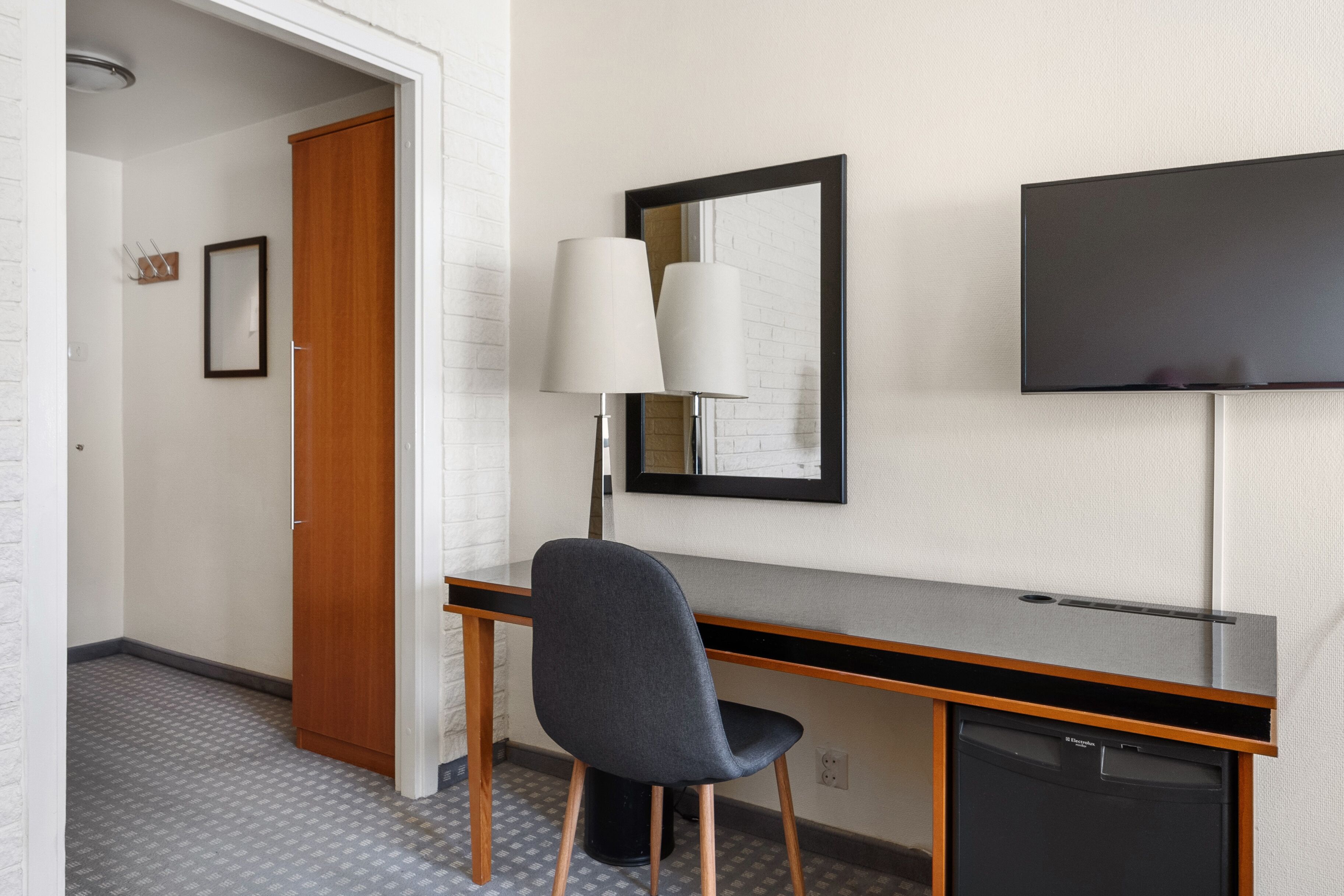 A hotel room with a wooden desk, a chair, a flat-screen TV mounted on the wall, and a door leading to another room.