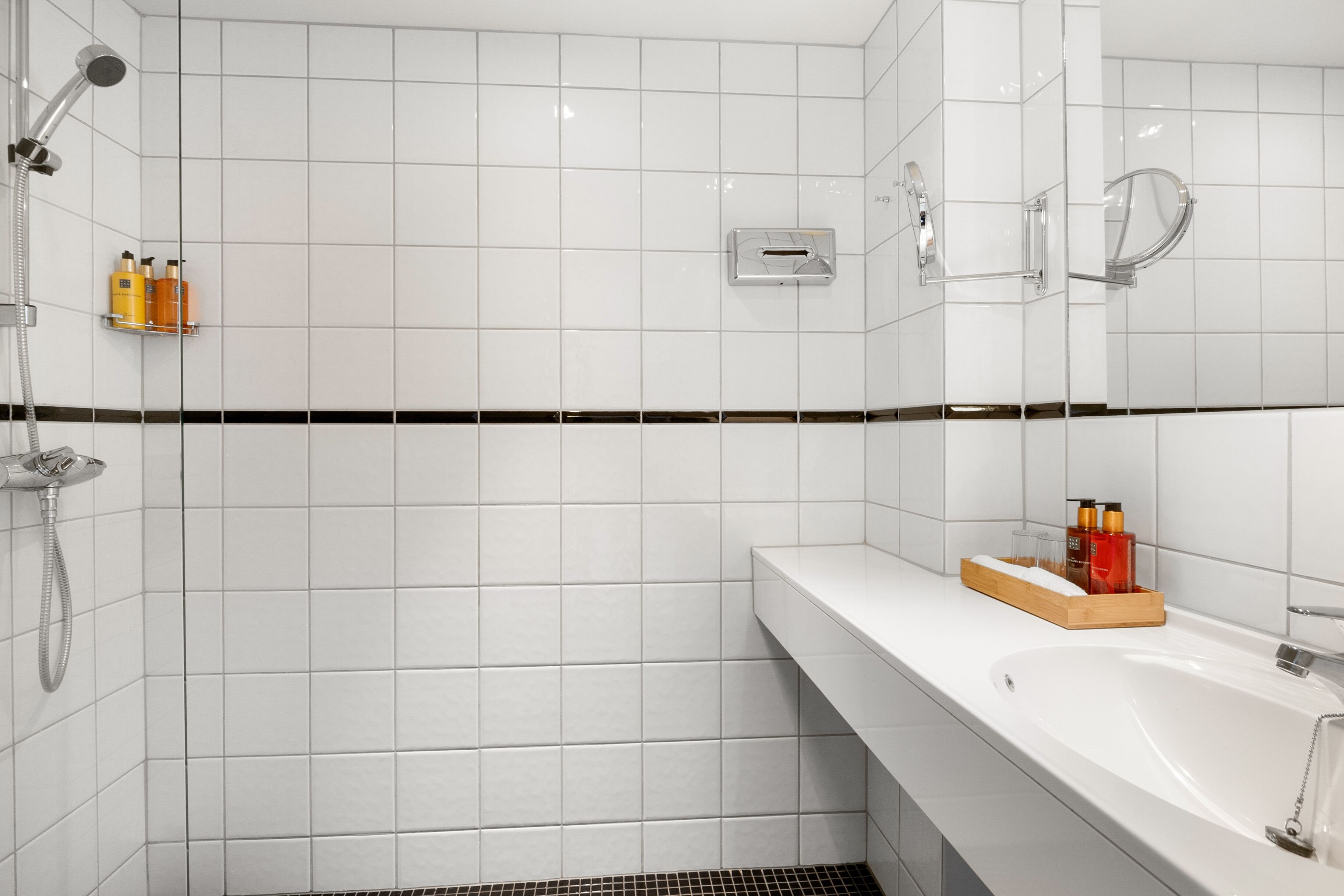 Un baño moderno con azulejos blancos, un lavamanos blanco y una ducha con cabezal de ducha manual.
