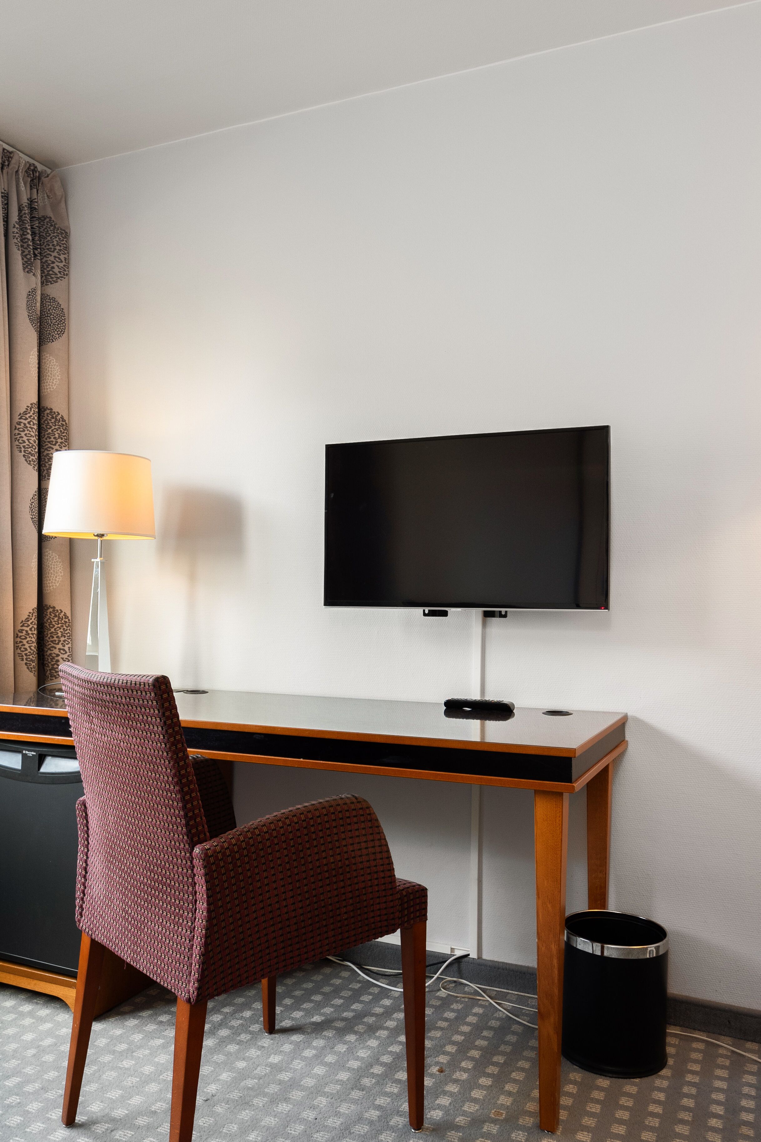 A hotel room with a desk, a television, a chair, a lamp, and a trash can.