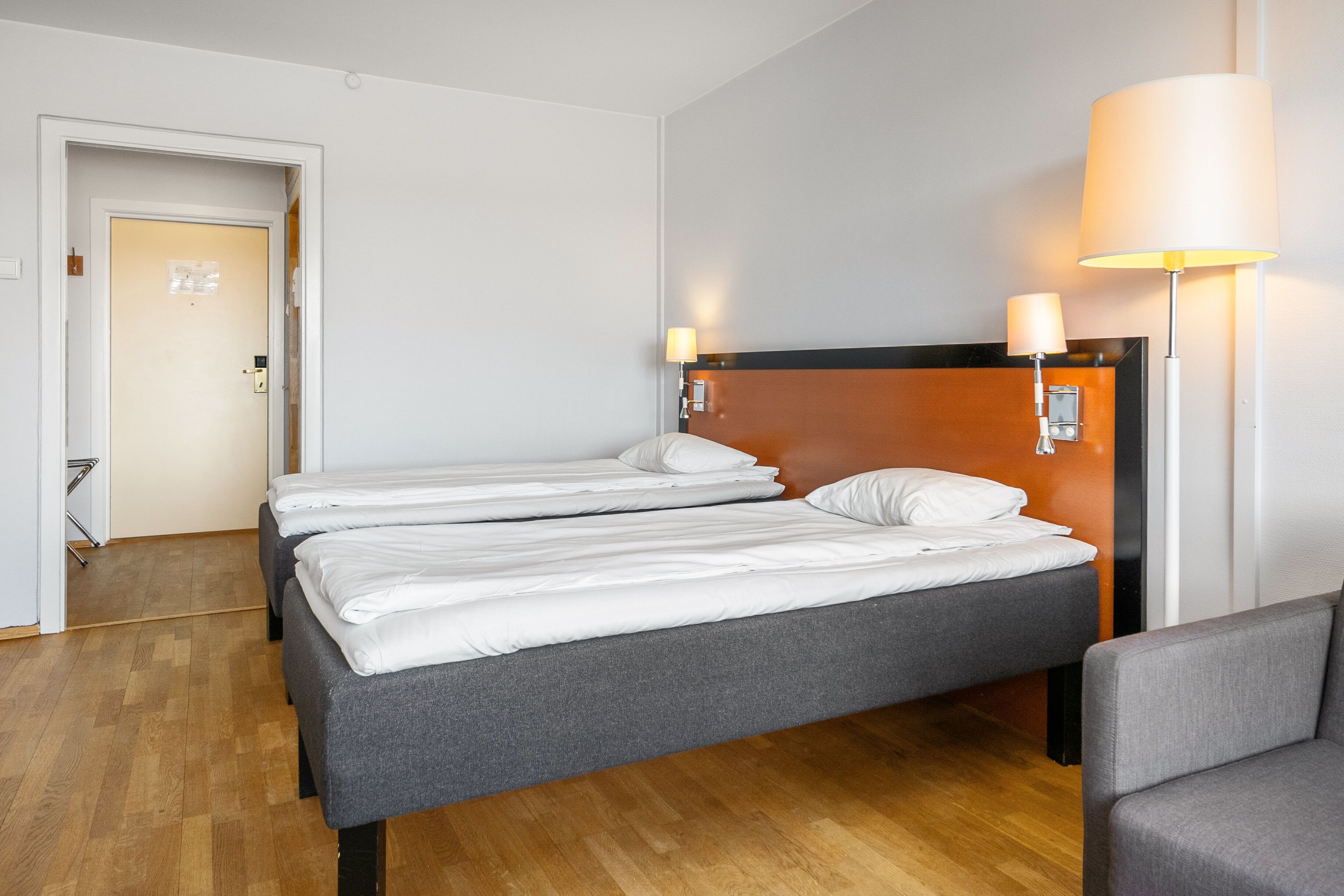 Habitación de hotel con dos camas individuales, suelo de madera, sofá gris y dos lámparas fijadas a la pared.