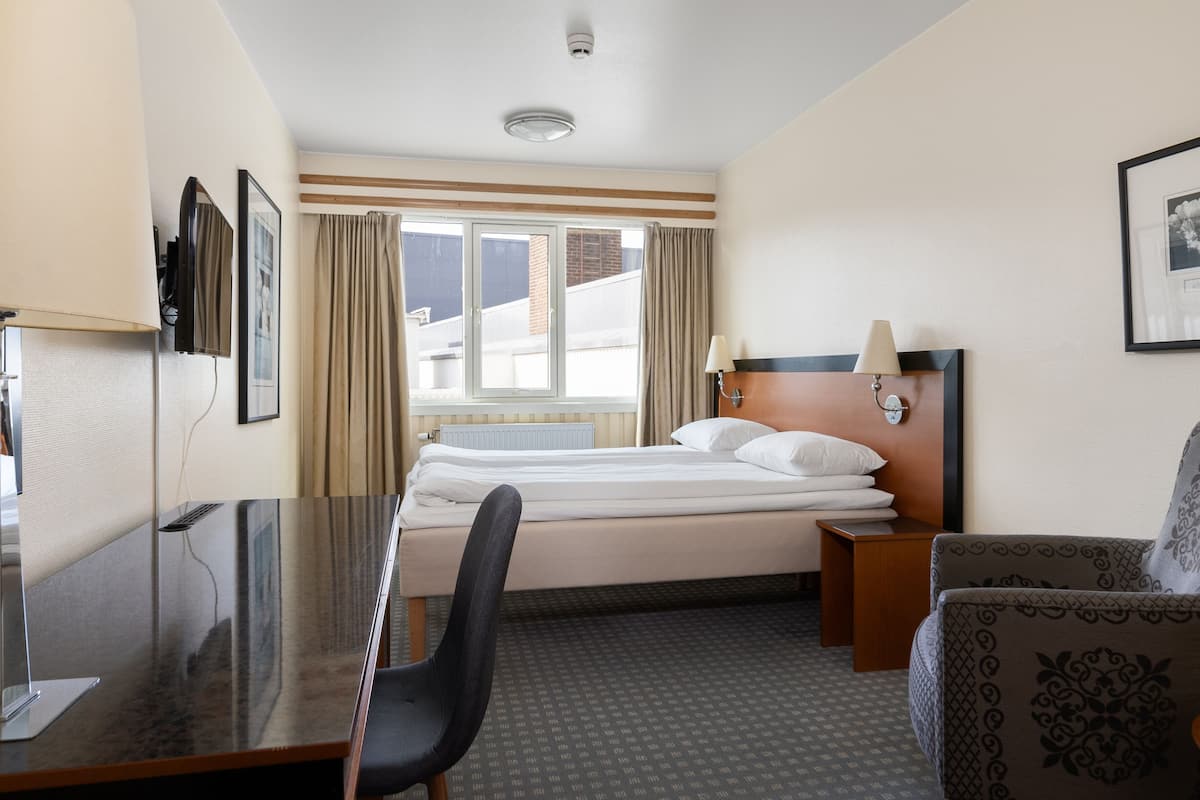 A hotel room with a bed, a desk, a chair, a lamp, and a window with curtains.