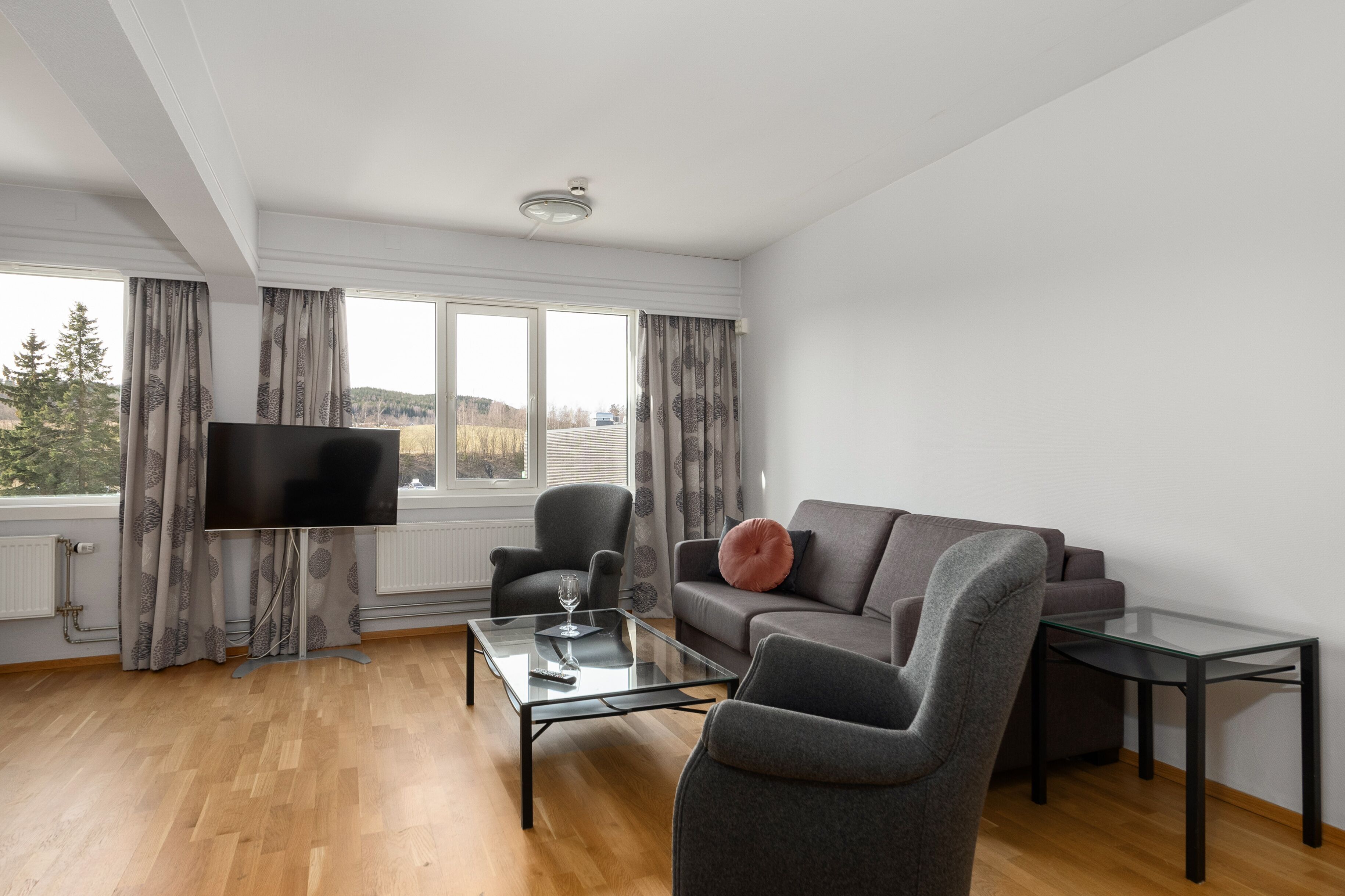 A modern living room with a flat-screen TV, grey sofa, armchairs, a glass coffee table, and a small side table.