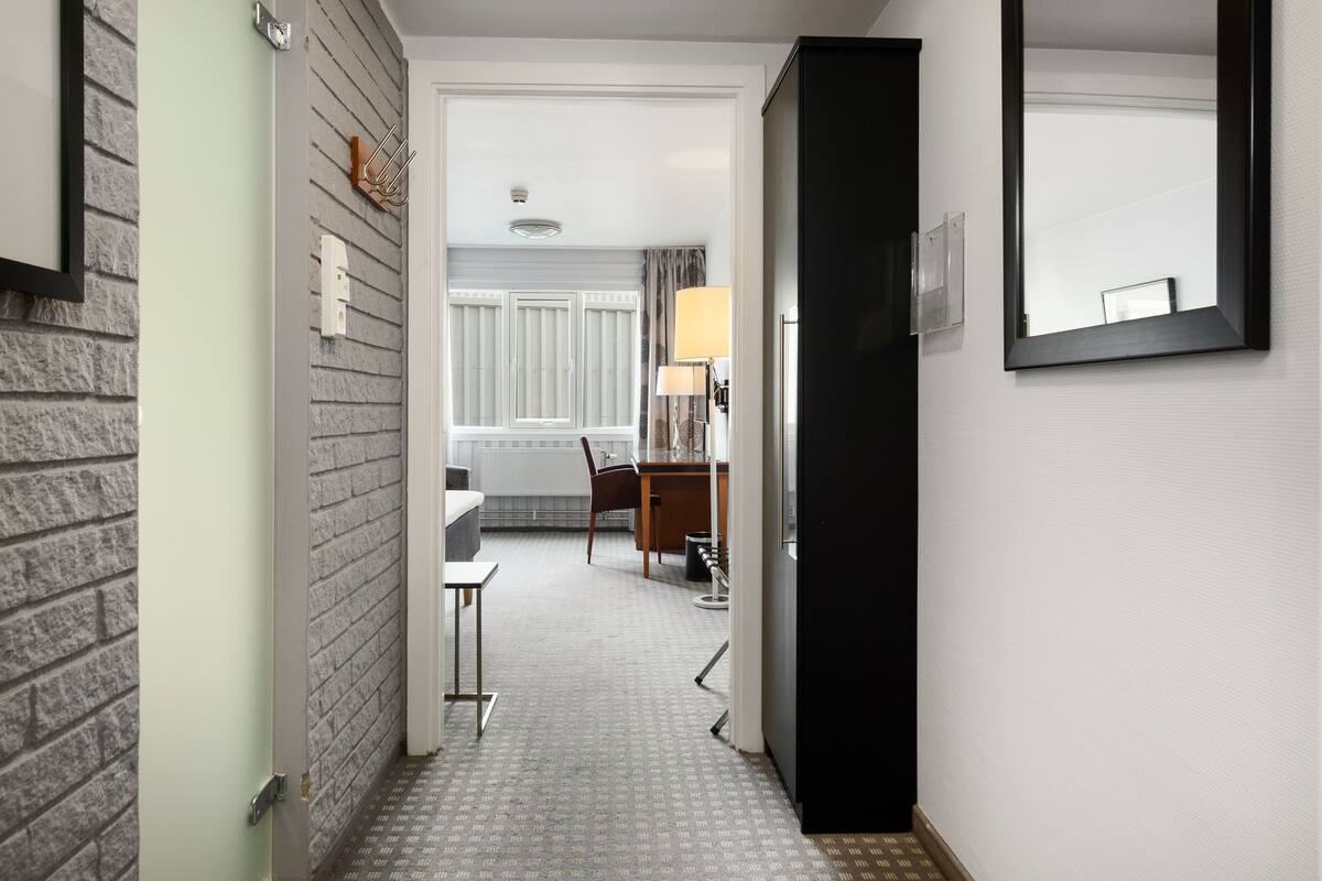 A narrow hallway with a brick wall on one side and a white wall on the other. A door is visible on the left, leading to a room with a bed and a window. A desk and chair are positioned in the center of the hallway.