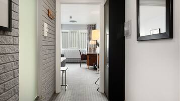 A narrow hallway with a brick wall on one side and a white wall on the other. A door is visible on the left, leading to a room with a bed and a window. A desk and chair are positioned in the center of the hallway.