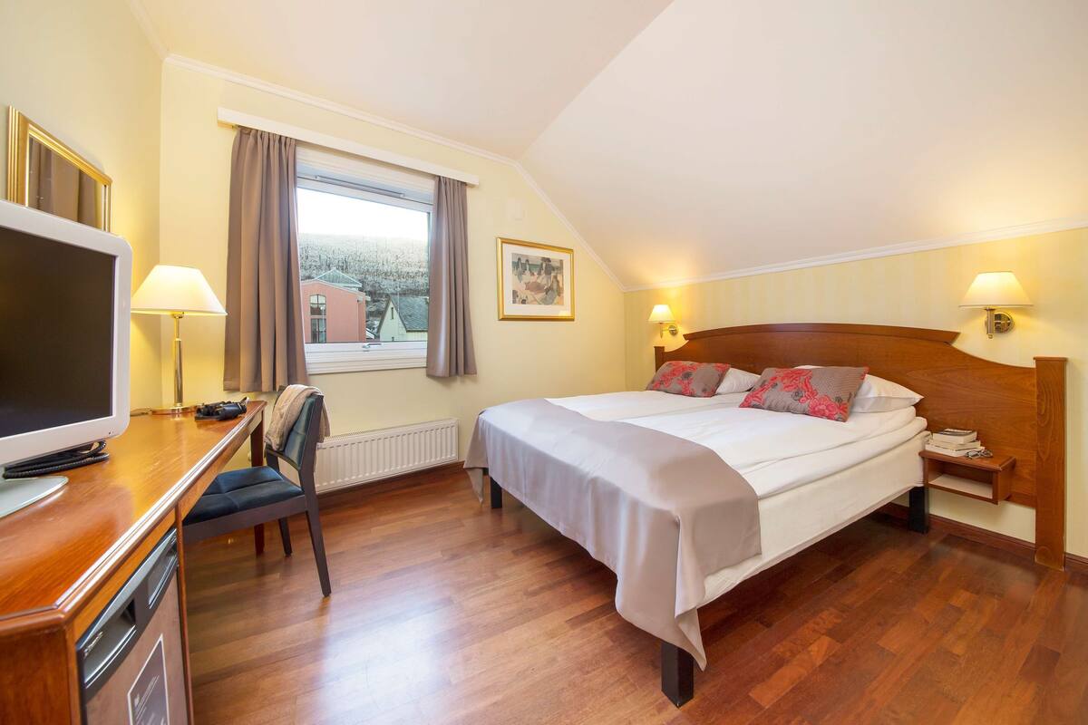 Ein Hotelzimmer mit Bett, Schreibtisch, Stuhl, Fernseher, Fenster mit Vorhängen und einem Bild an der Wand.