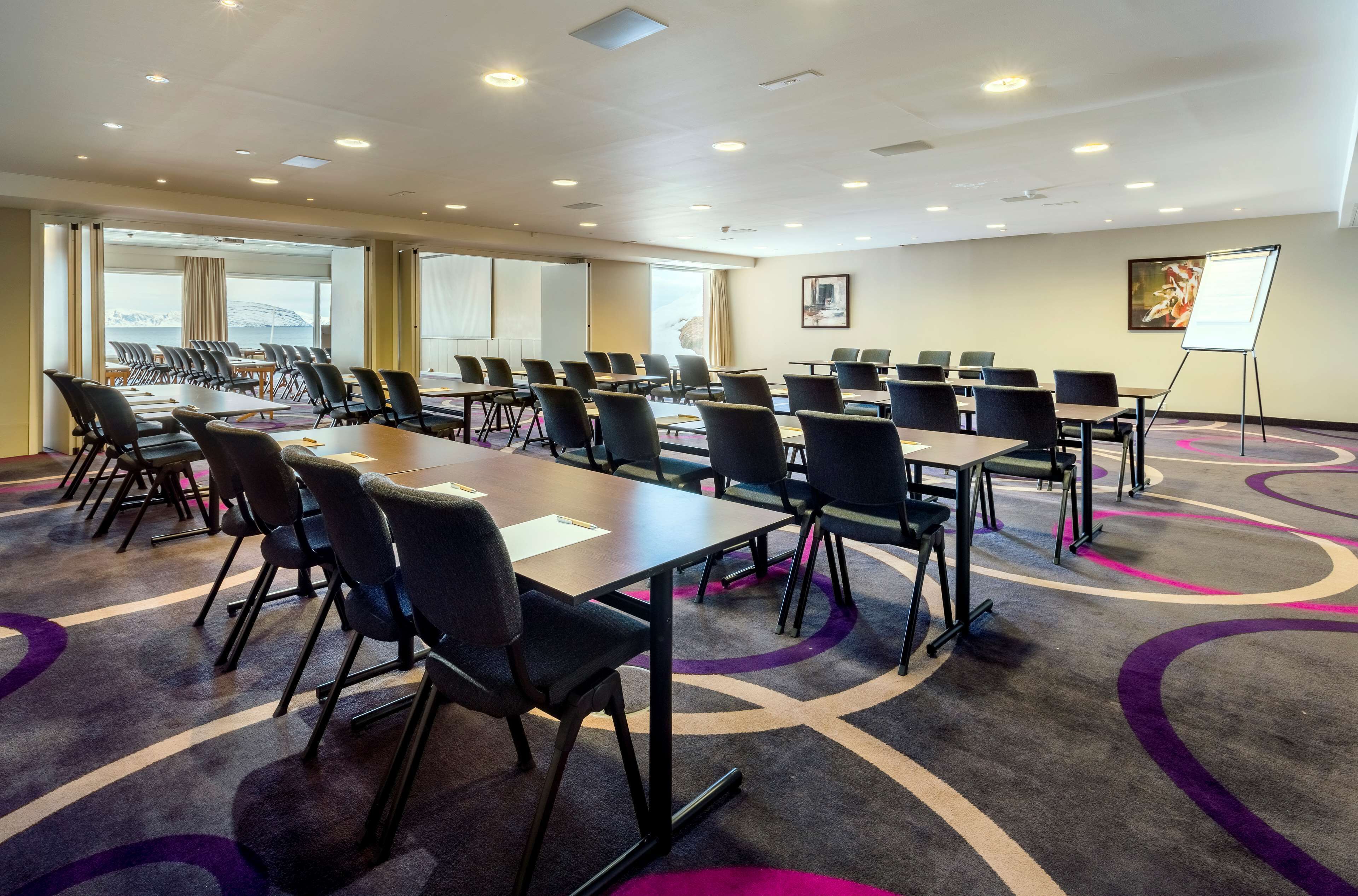 Uma sala de conferências com várias mesas e cadeiras, janelas amplas e uma tela de projeção.