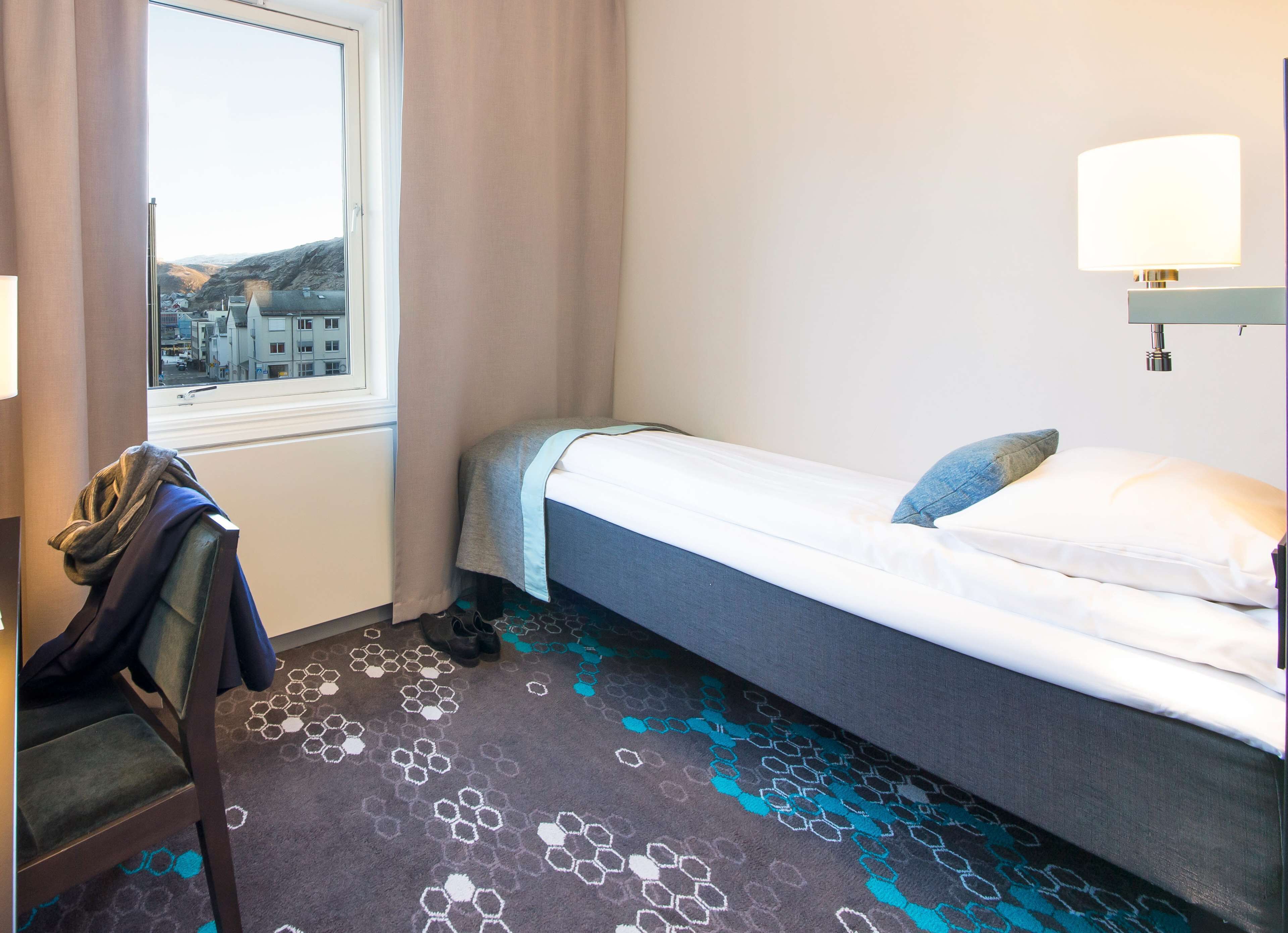 Habitación de hotel con cama, una silla, ventana y vistas a edificios, además de una lámpara.