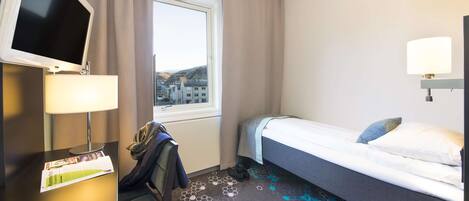Habitación de hotel con una cama, un escritorio con televisor, una lámpara y una ventana con cortinas.