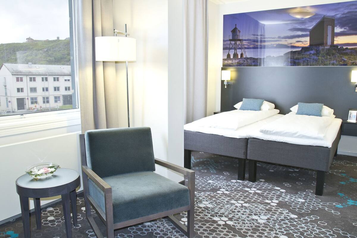 Ein Hotelzimmer mit einem Bett, einem Sessel, einem kleinen Tisch, einer Lampe und Blick auf Gebäude durch das Fenster.
