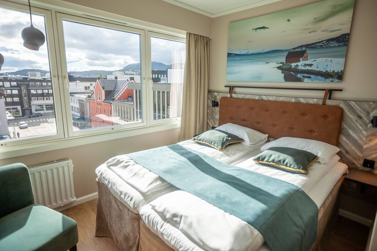 A hotel room with a bed, a view of buildings, a radiator, and a window.