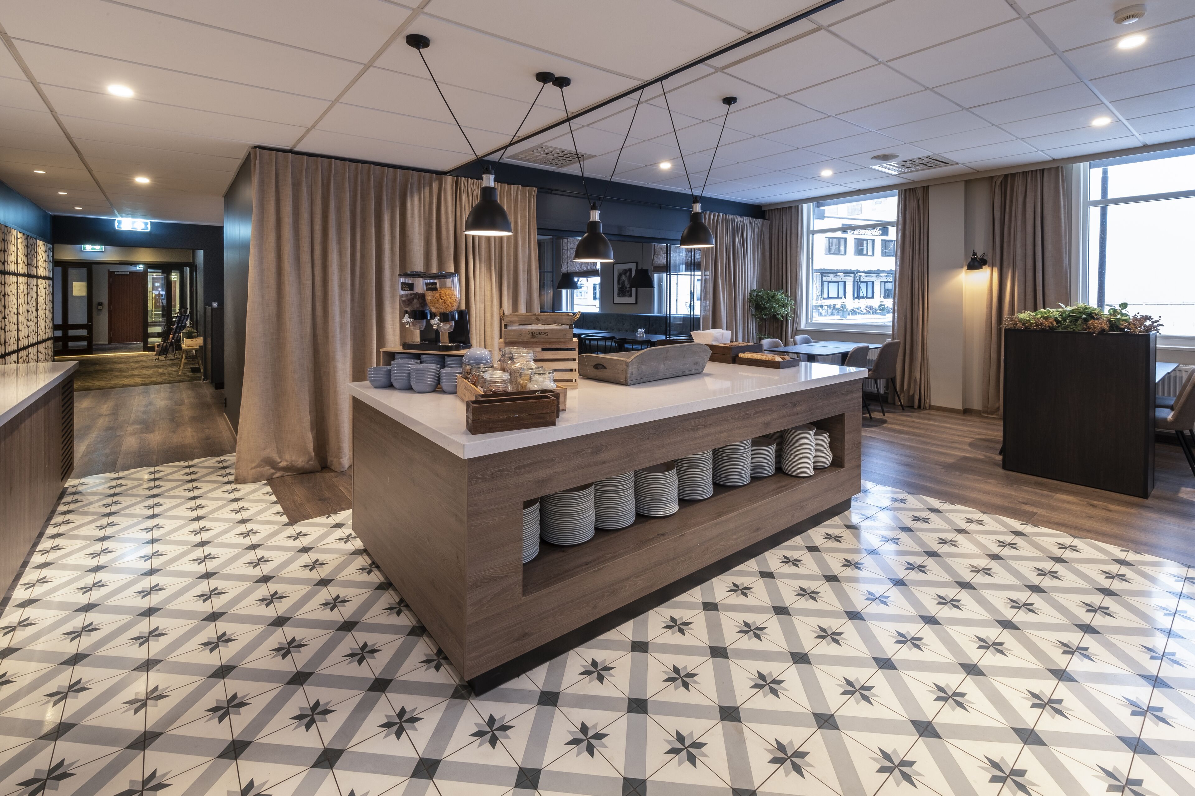 A modern hotel lobby with a central bar area, patterned floor tiles, and large windows.