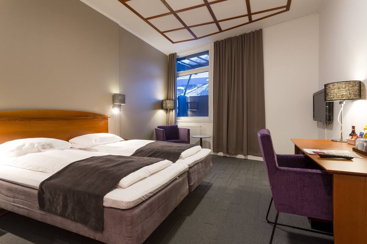 A hotel room with a bed, a desk, a chair, a TV, and a window with curtains.