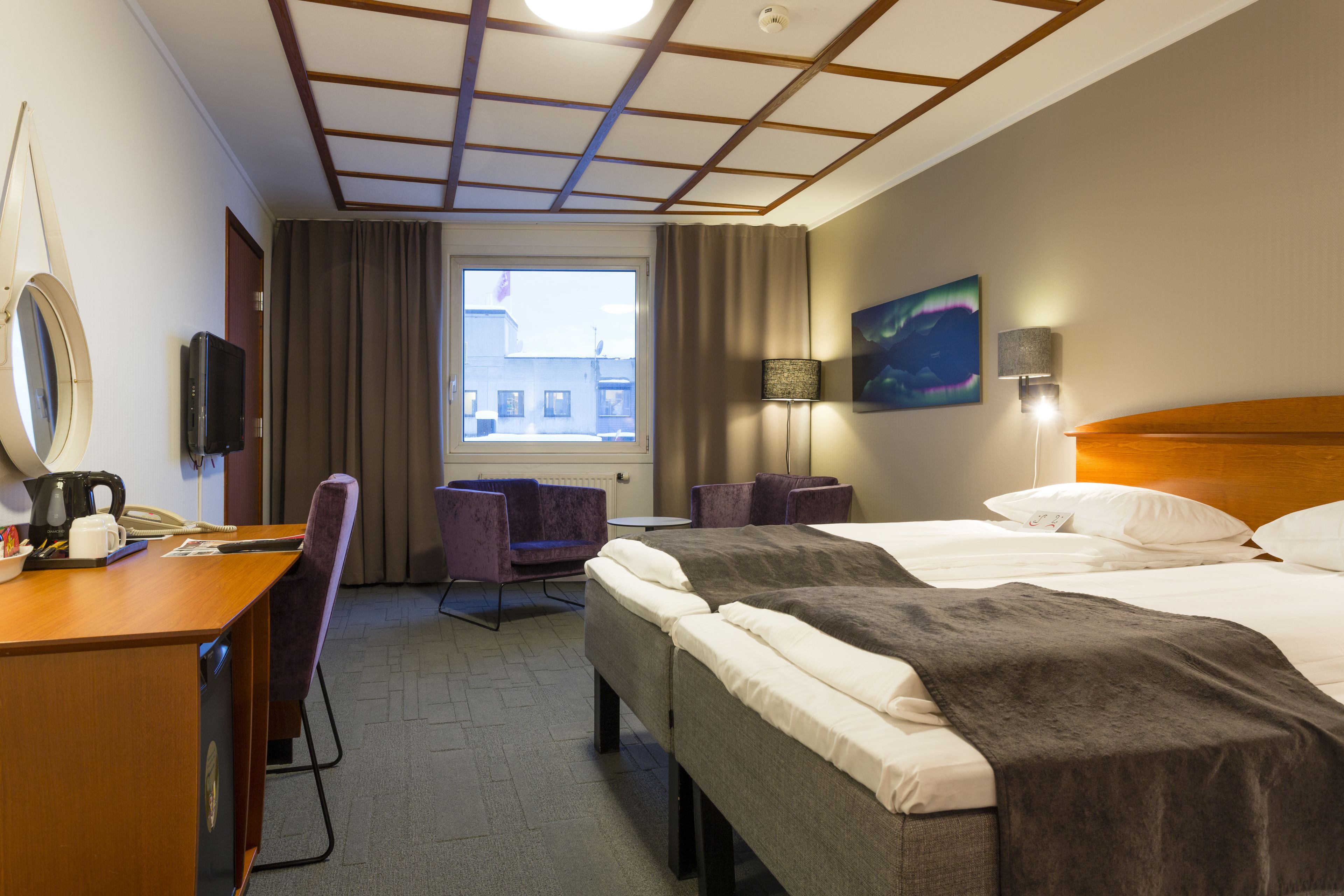 A hotel room with a wooden desk, two beds, a chair, a window, a painting, and a lamp.