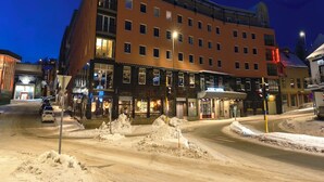 A multi-story hotel with a modern design, located at a snowy intersection with visible snowbanks and a car in the foreground. - Scandic Harstad (Harstad)