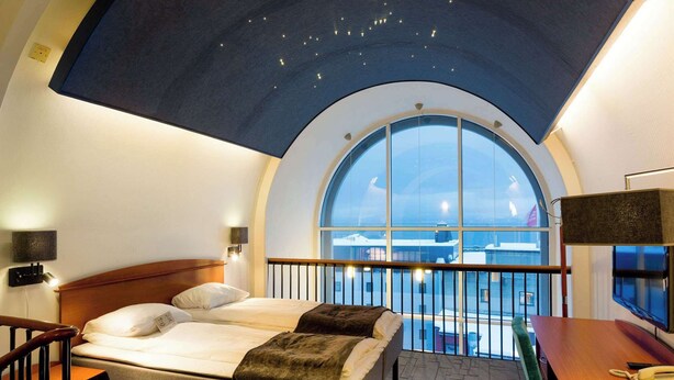 A hotel room with a large arched window, a bed with a view of the sea, a desk with a telephone, and a ceiling with a starry design. - Scandic Harstad (Harstad)