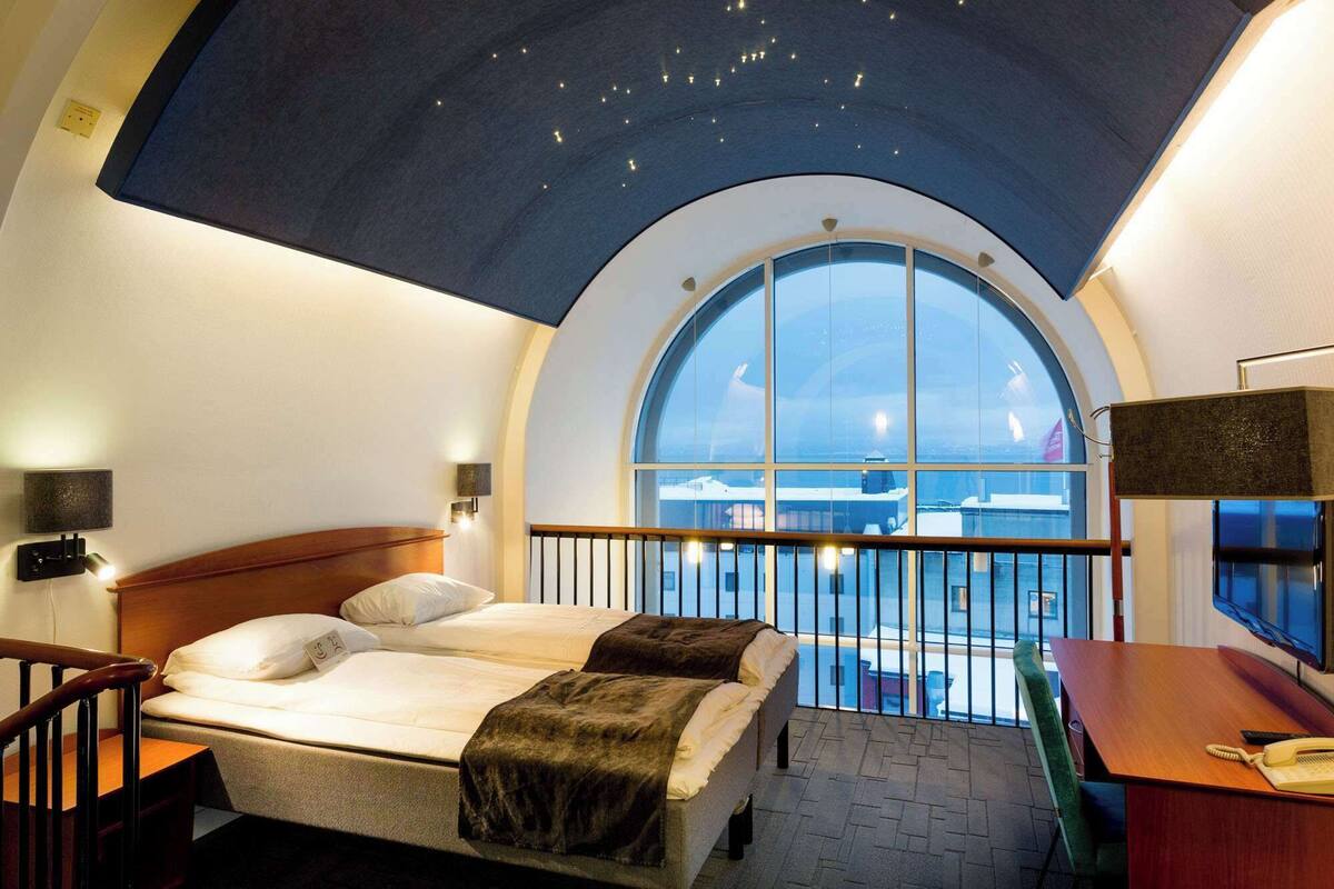 A hotel room with a large arched window, a bed with a view of the sea, a desk with a telephone, and a ceiling with a starry design.