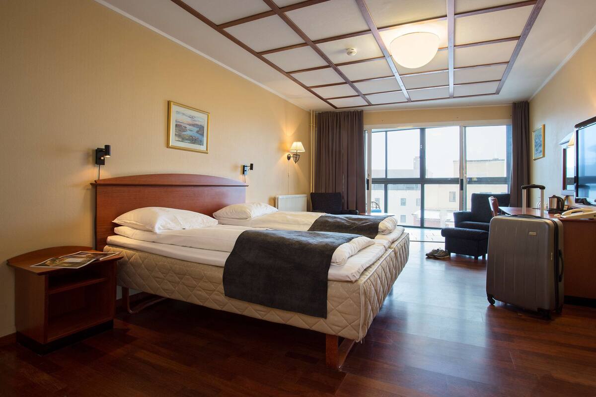 A hotel room with two beds, a desk, a chair, a suitcase, and a large window.