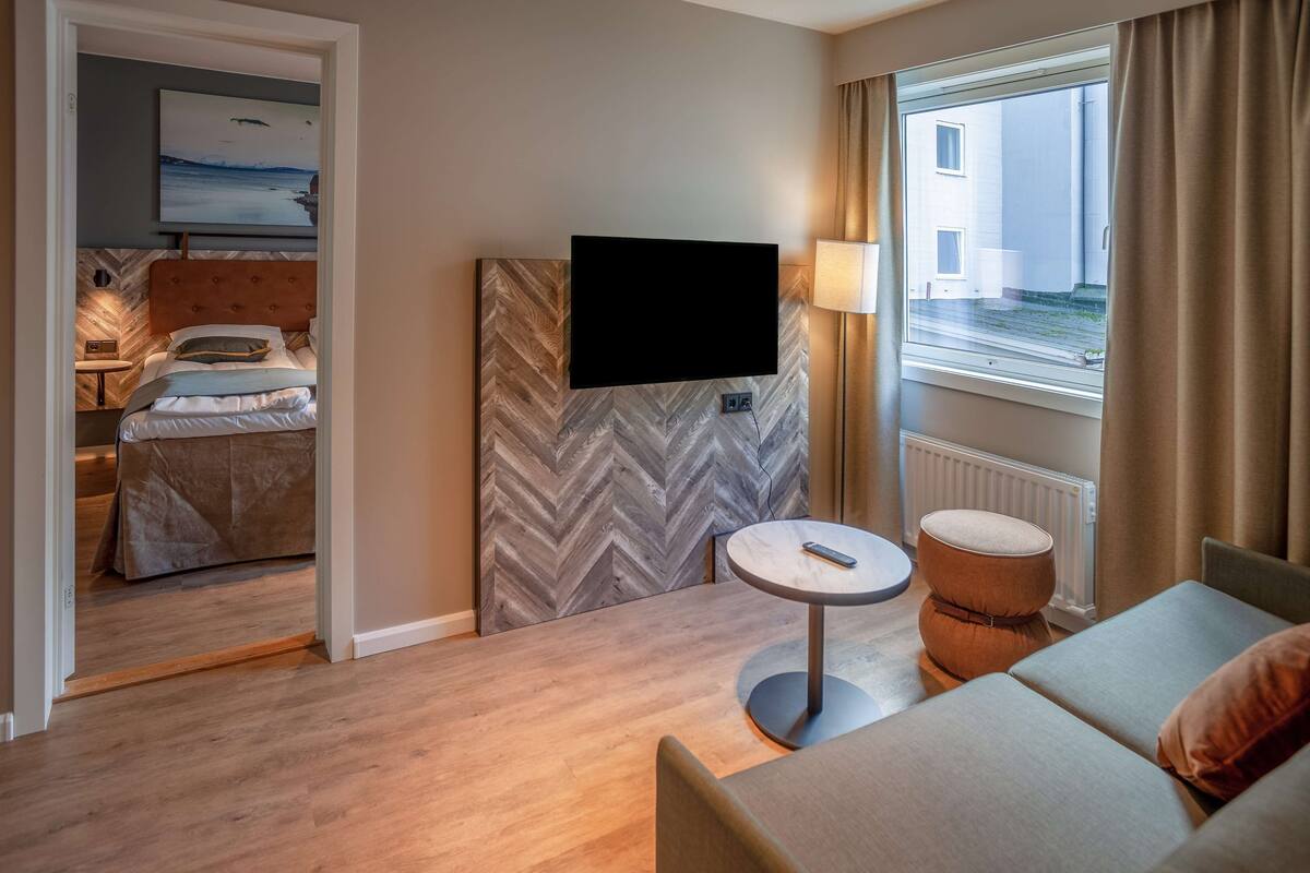 A hotel room with a bed, a TV mounted on a herringbone-patterned wall, a round table with a remote, a cushioned armchair, and a view of a building through a window.