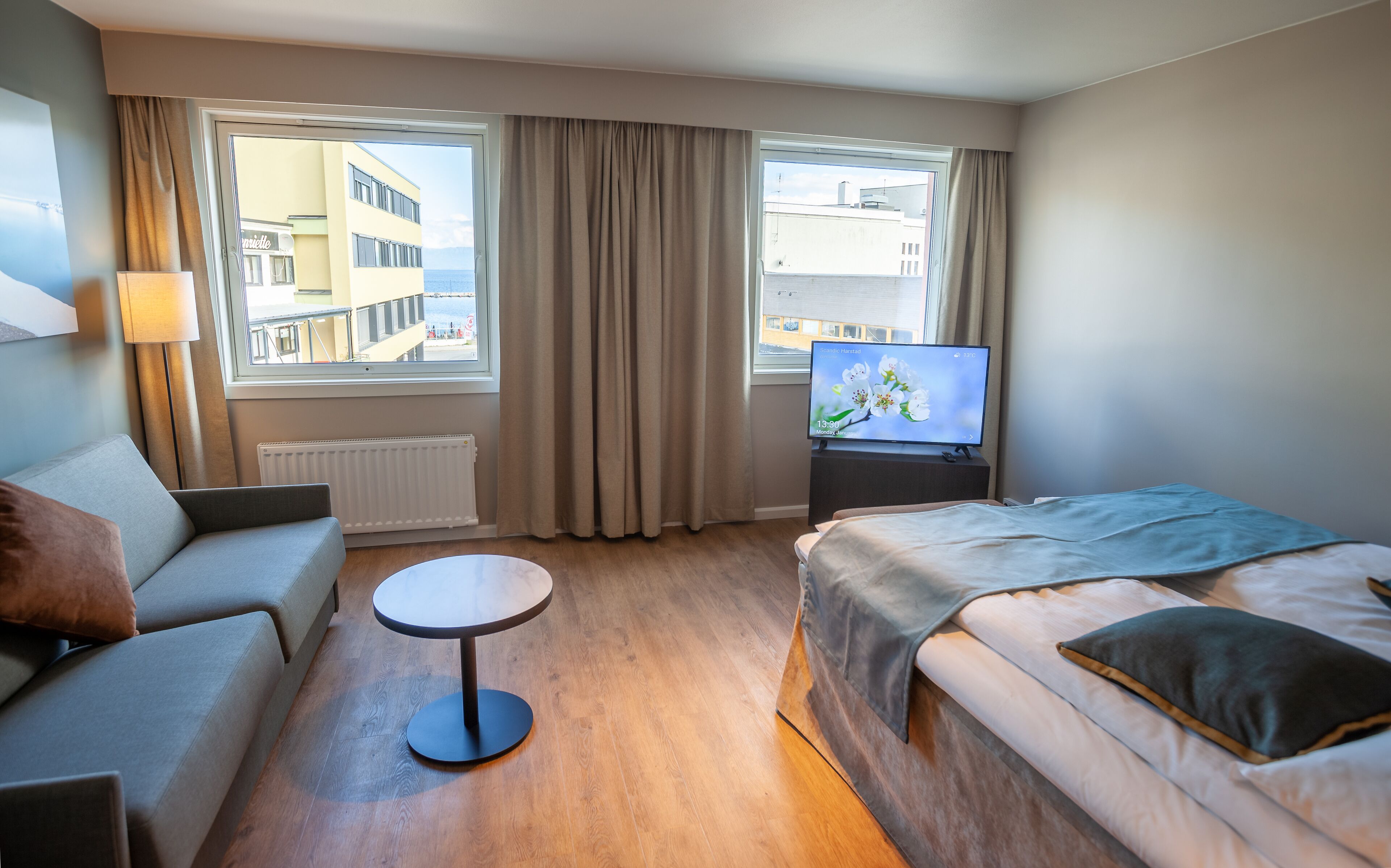 A hotel room with a bed, a sofa, a small table, a TV, and large windows with curtains.