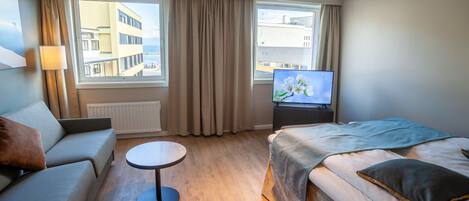 A hotel room with a bed, a sofa, a small table, a TV, and large windows with curtains.