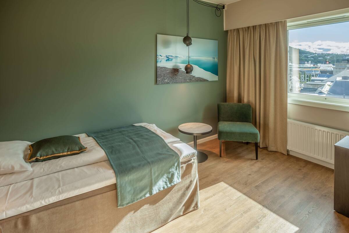 A hotel room with a bed, a chair, a small table, a painting, and a window with a view of a cityscape.
