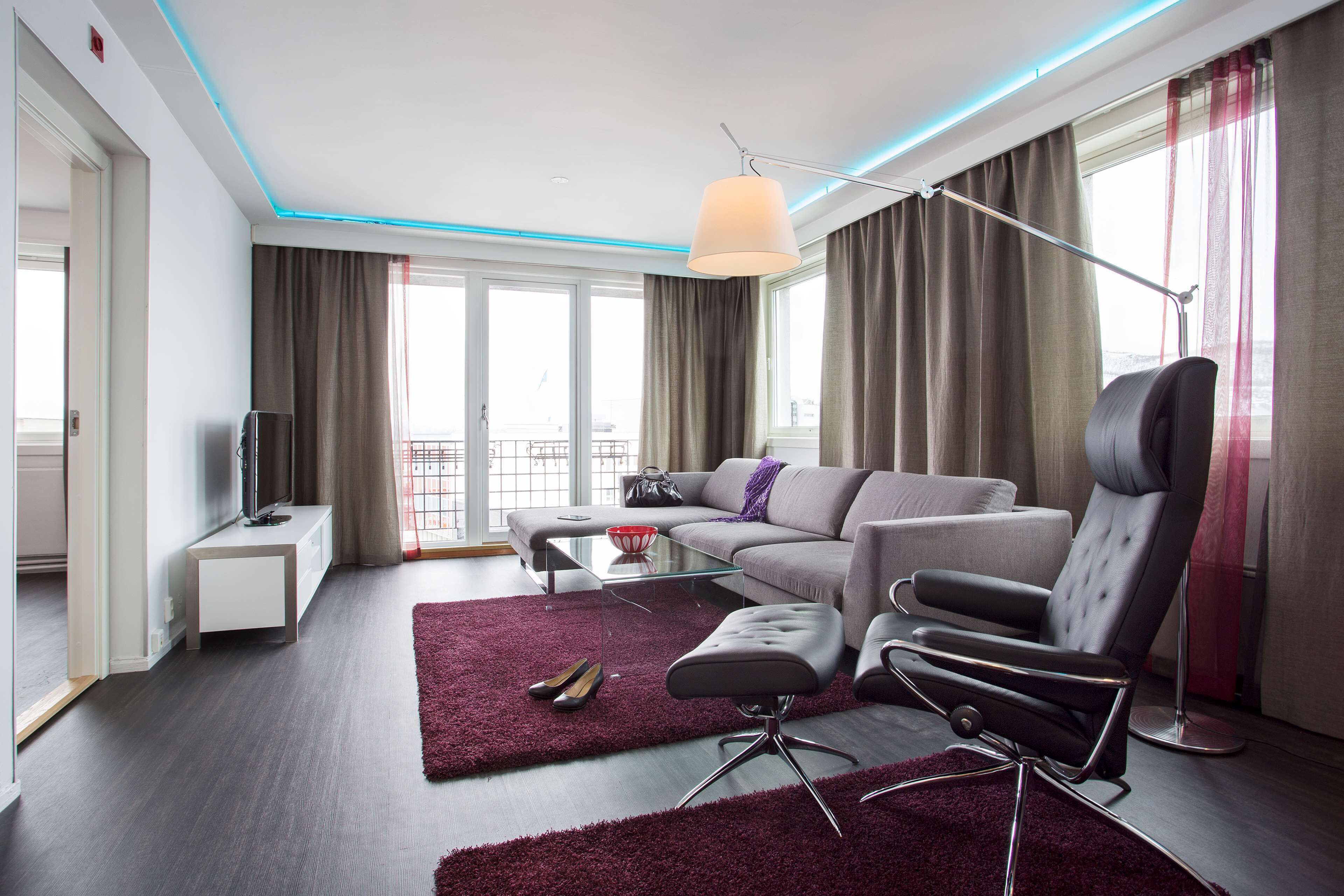 A modern living room with a sofa, ottoman, and a recliner, a glass coffee table, a flat-screen TV, and a large window with curtains.