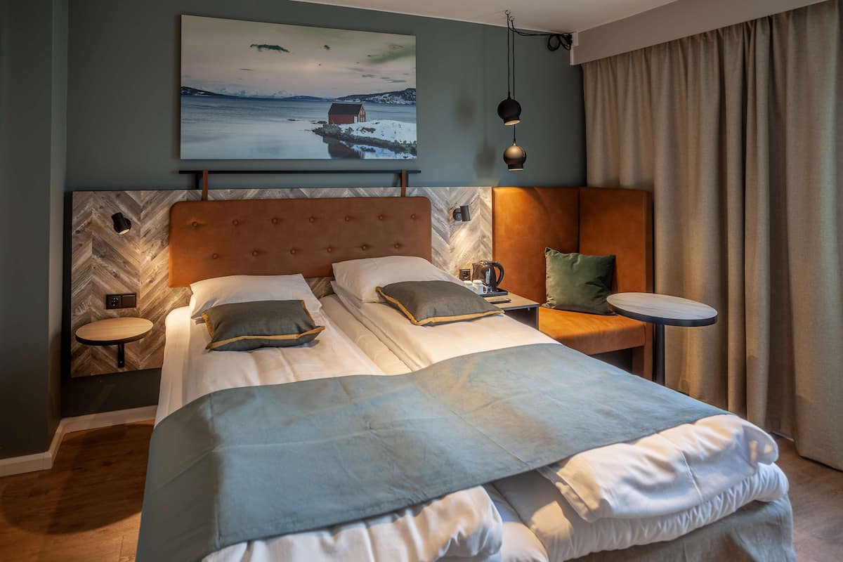 A modern hotel room with a large bed, a small bedside table, a nightstand, and a wall-mounted projector displaying a scenic image.