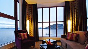 Una habitación con un ventanal grande, un sillón morado, un sofá y una mesa con una lámpara.