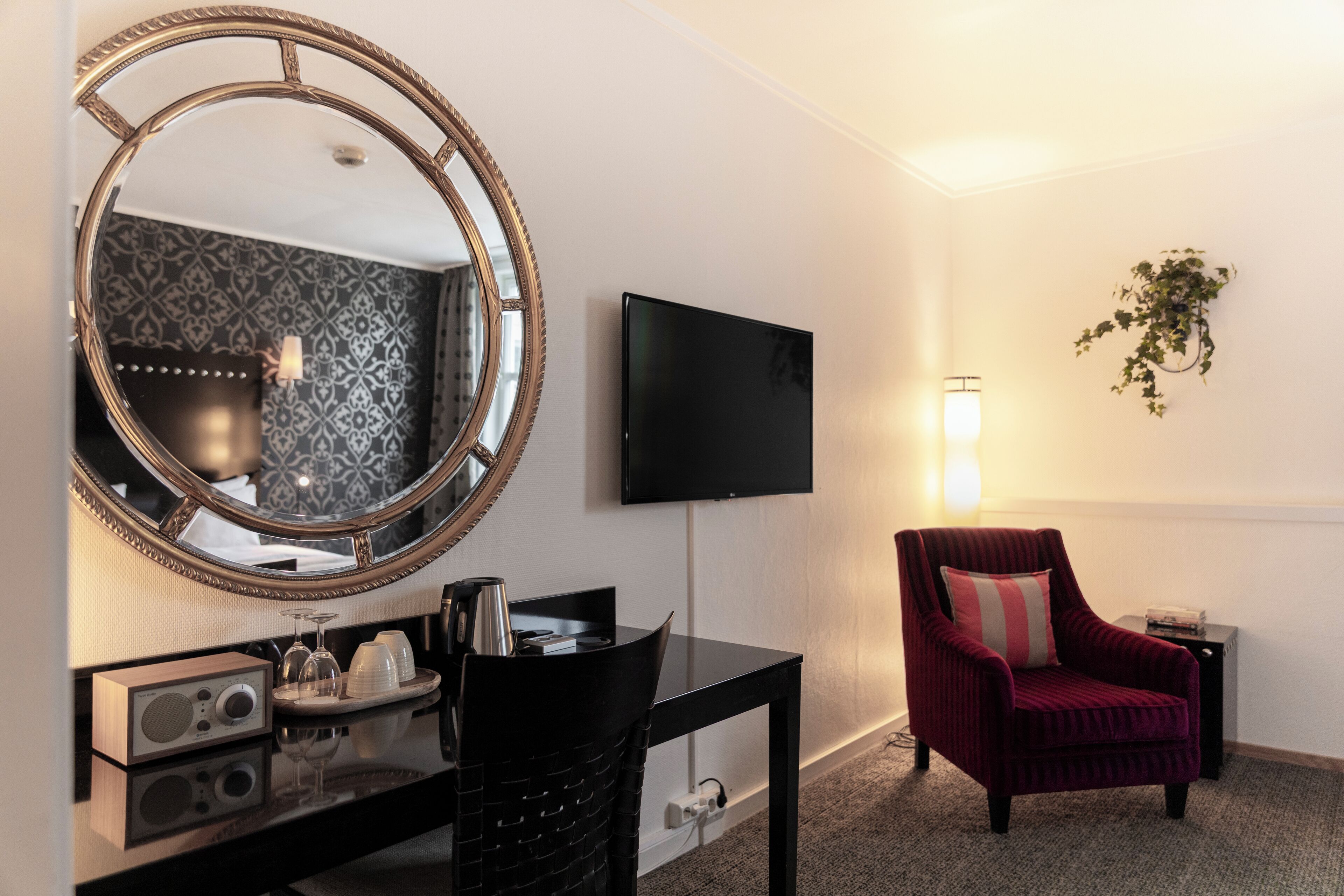 Ein Hotelzimmer mit einem runden Spiegel, einem Flachbildfernseher, einem roten Sessel, einem kleinen Tisch mit einem Radio und einer Pflanze an der Wand.