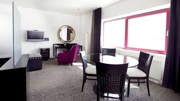 Ett hotellrum med ett runt glasbord, fyra svarta stolar, en tv, en spegel, ett litet skrivbord och ett fönster med gardiner.