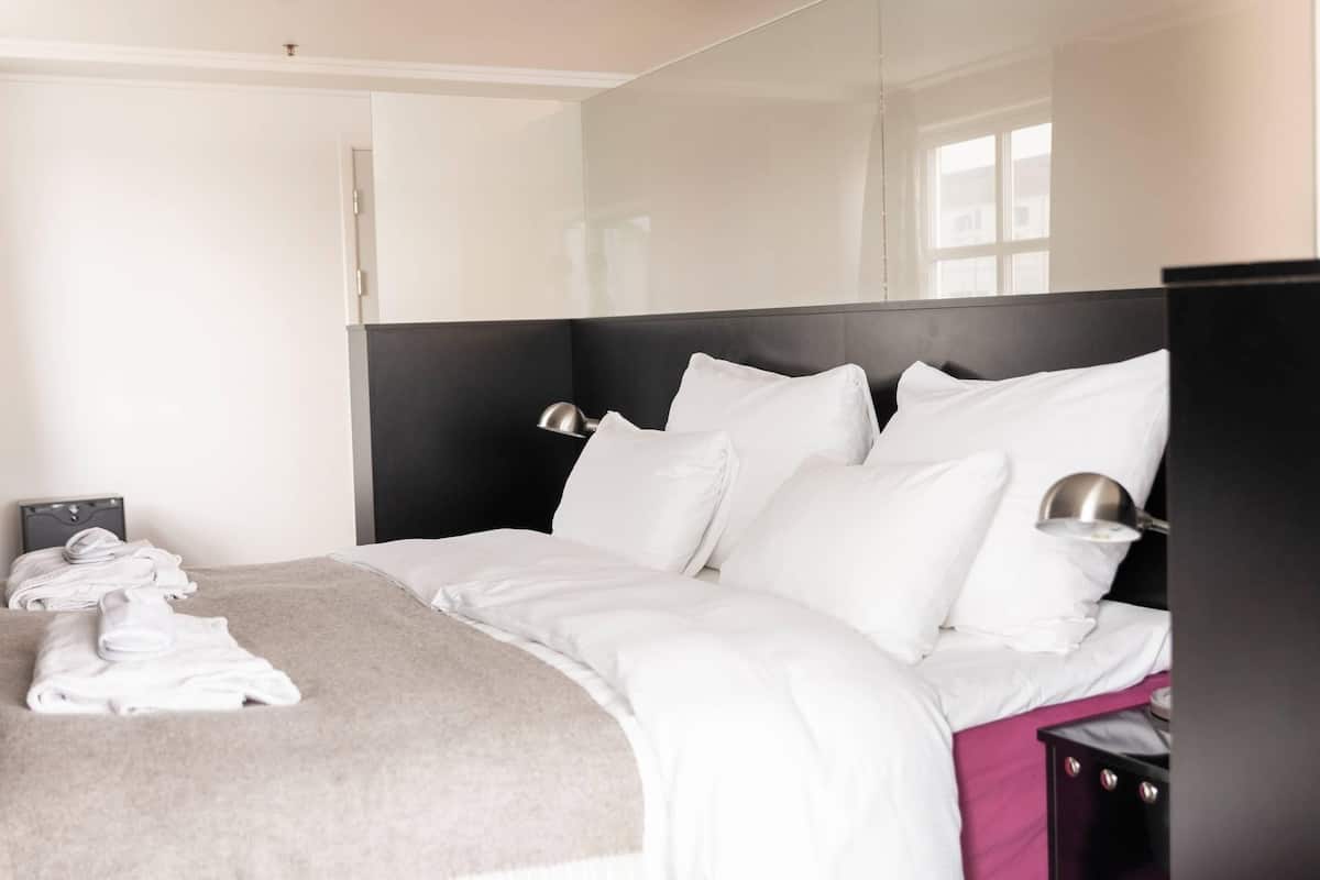 Una cama bien hecha con sábanas blancas y una manta gris, una mesita de noche con lámpara y un espejo en la pared.