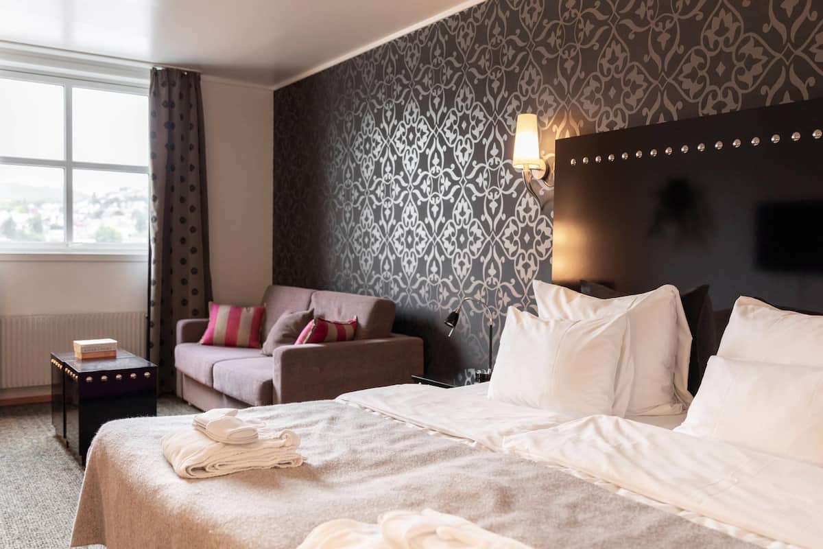 Habitación de hotel con cama, sofá, una mesita con un libro, lámpara y pared con diseño.