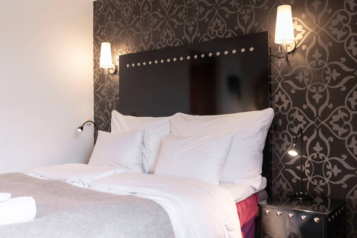 Una cama bien tendida con sábanas blancas, cabecero oscuro con tachuelas decorativas y dos lámparas fijadas a la pared.