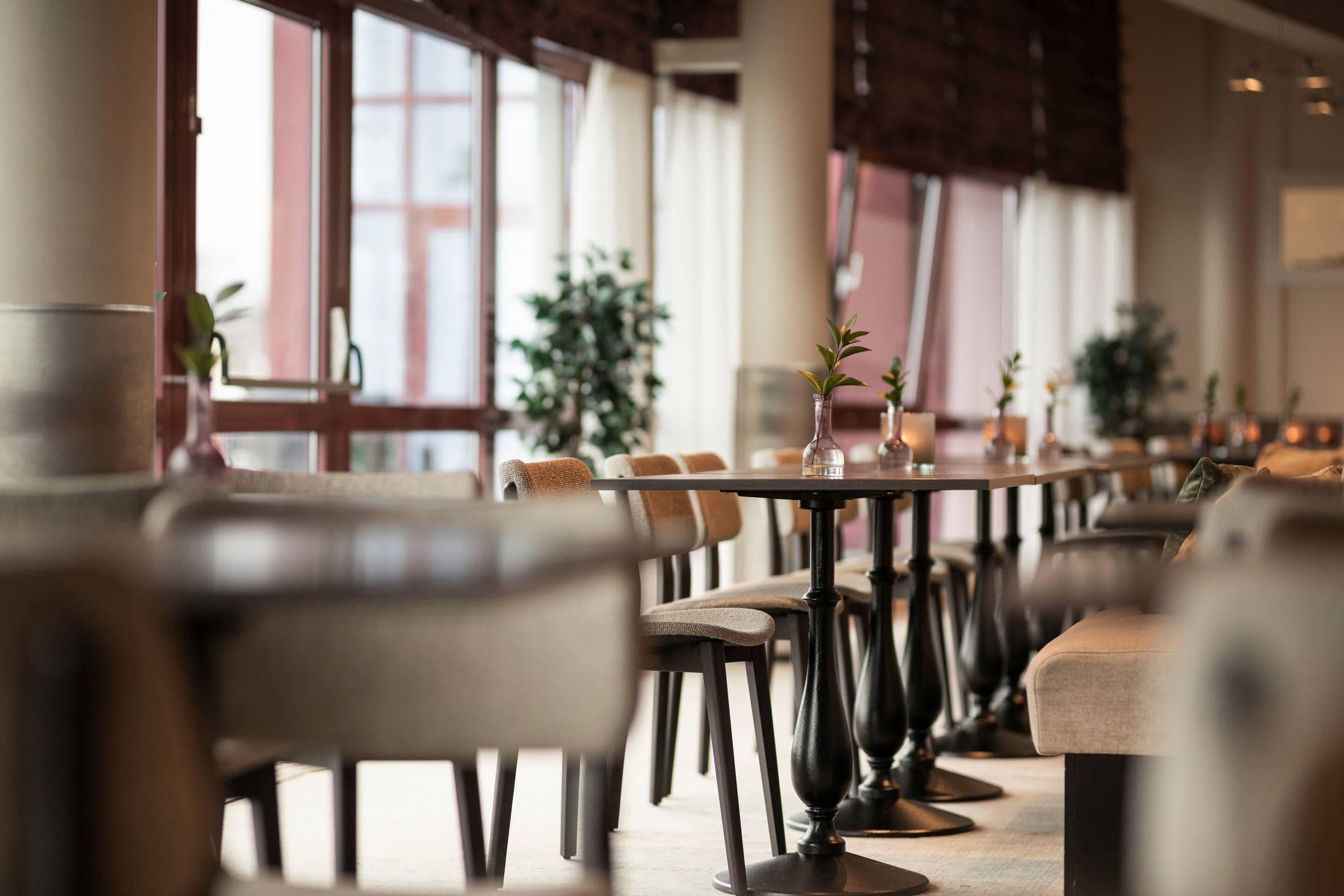 A dining area with tables and chairs, large windows, and potted plants.