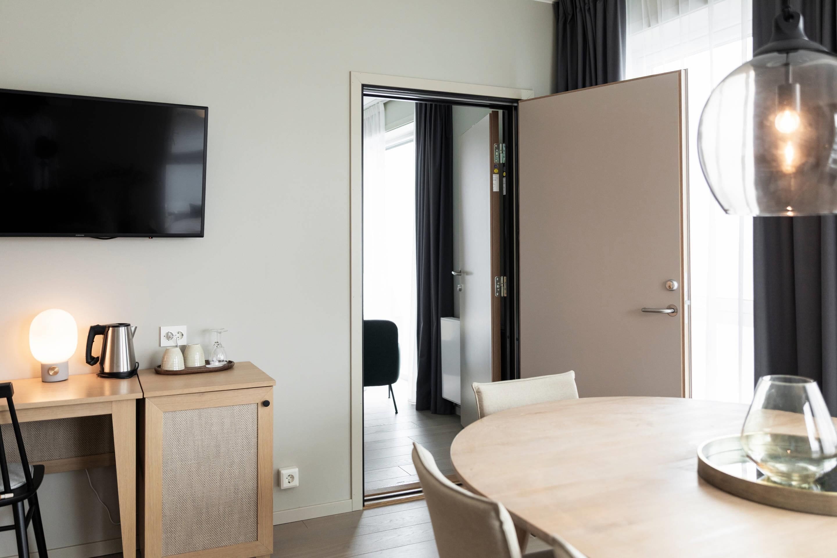 A modern hotel room with a flat-screen TV, a wooden desk with a kettle and cups, a round dining table with a glass, and a large mirror.