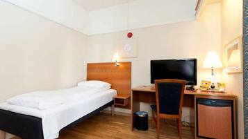 Ein Hotelzimmer mit einem Einzelbett, einem Schreibtisch mit Computer und Fernseher, einem Sessel und einer kleinen Mülleimer.