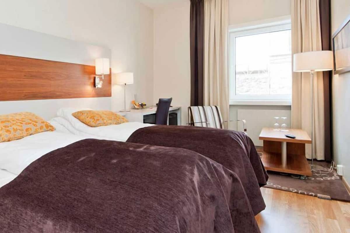 A hotel room with two beds, a desk, a chair, a lamp, and a view of the outdoors.