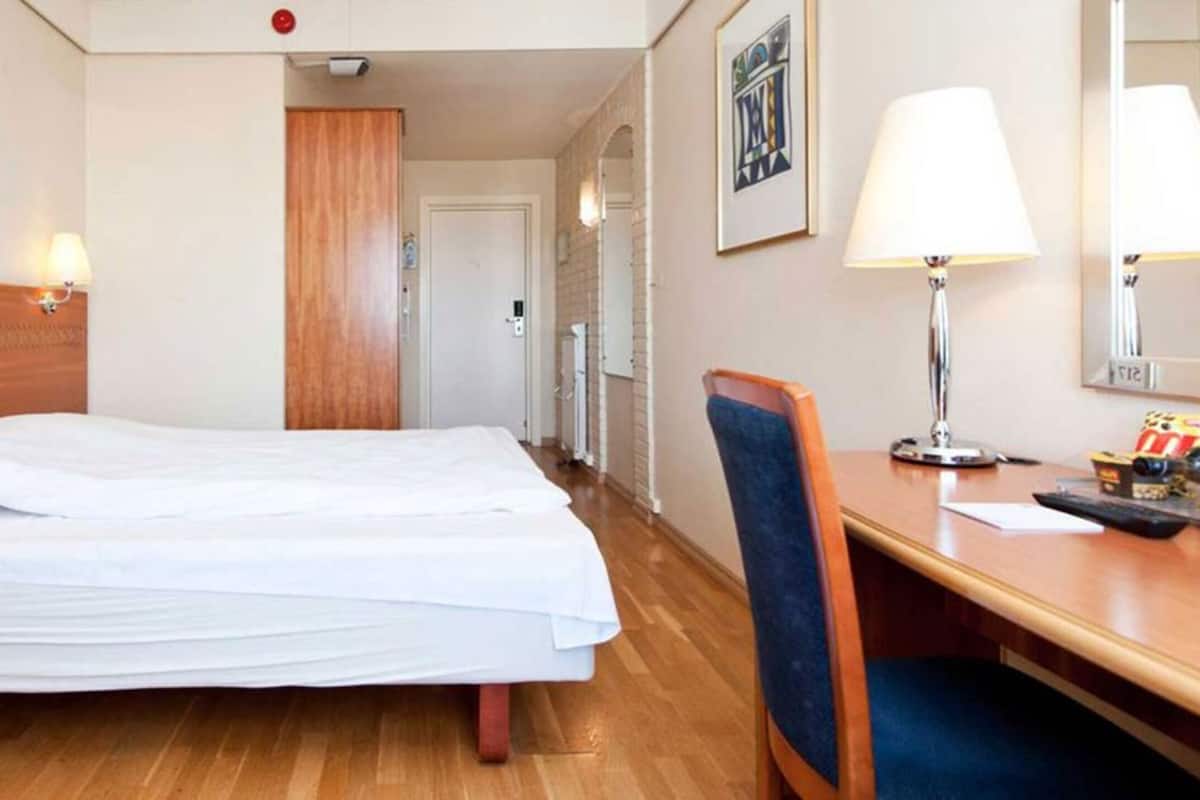 A hotel room with a double bed, wooden headboard, bedside lamps, a desk with a chair, and a mirror.