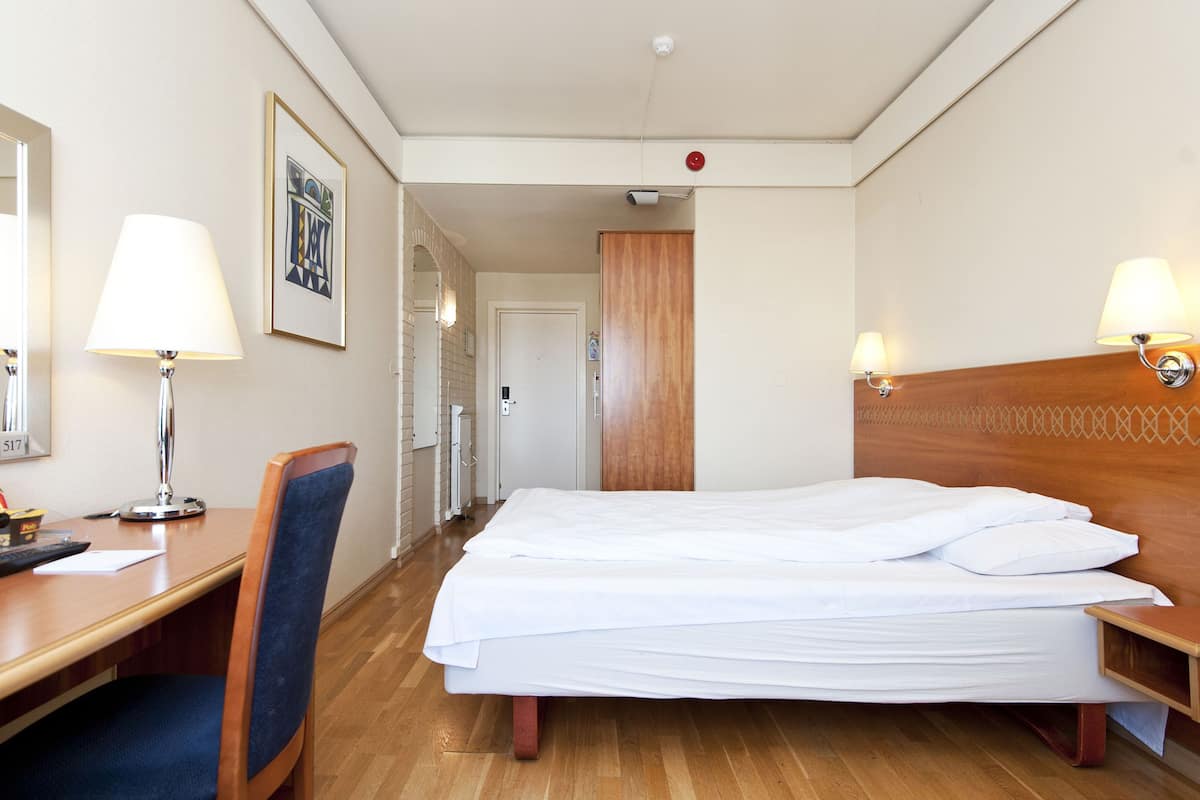 A hotel room with a bed, a desk with a chair, a mirror, and two wall-mounted lamps.