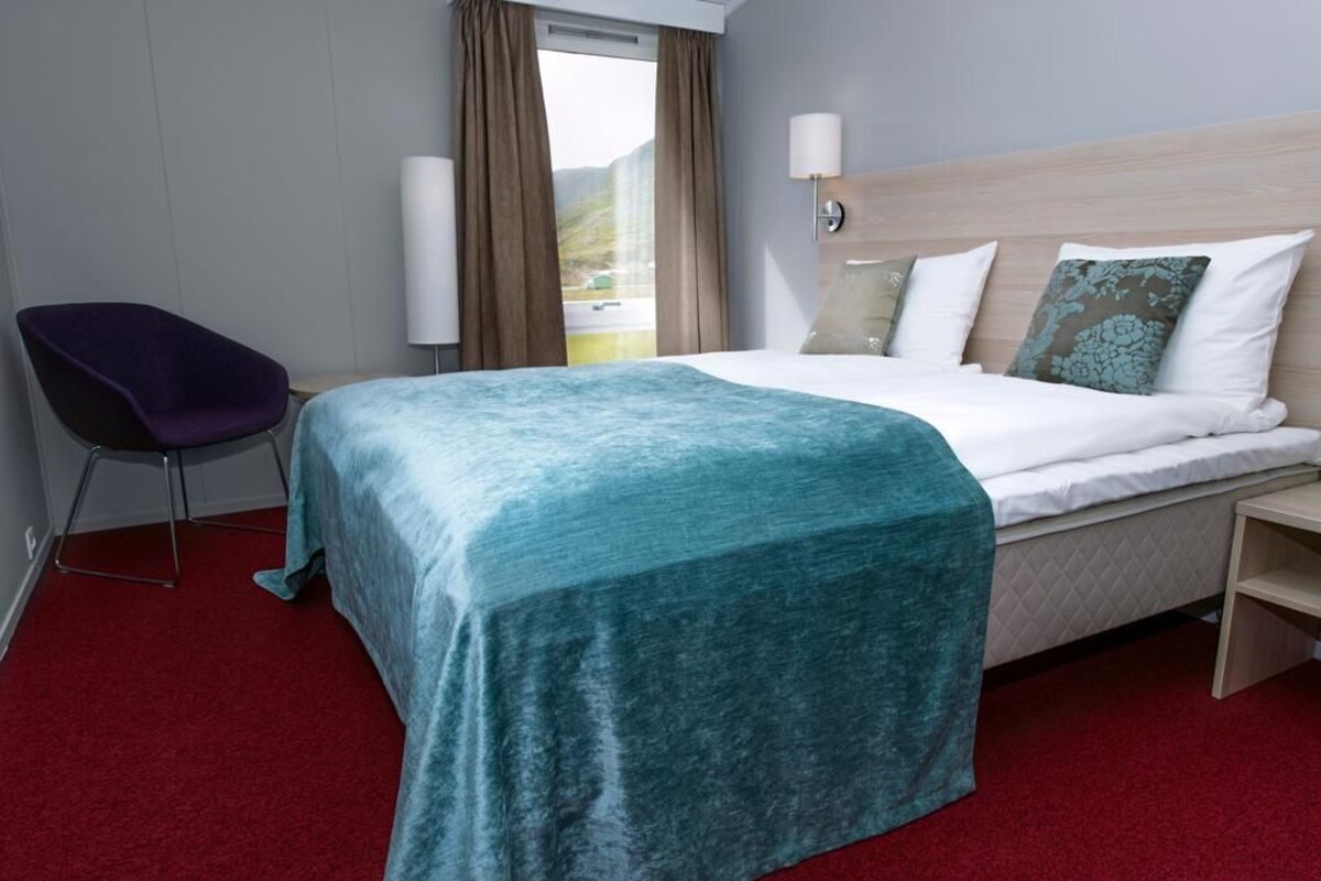 A hotel room with a bed, a chair, a nightstand, and a window with curtains.