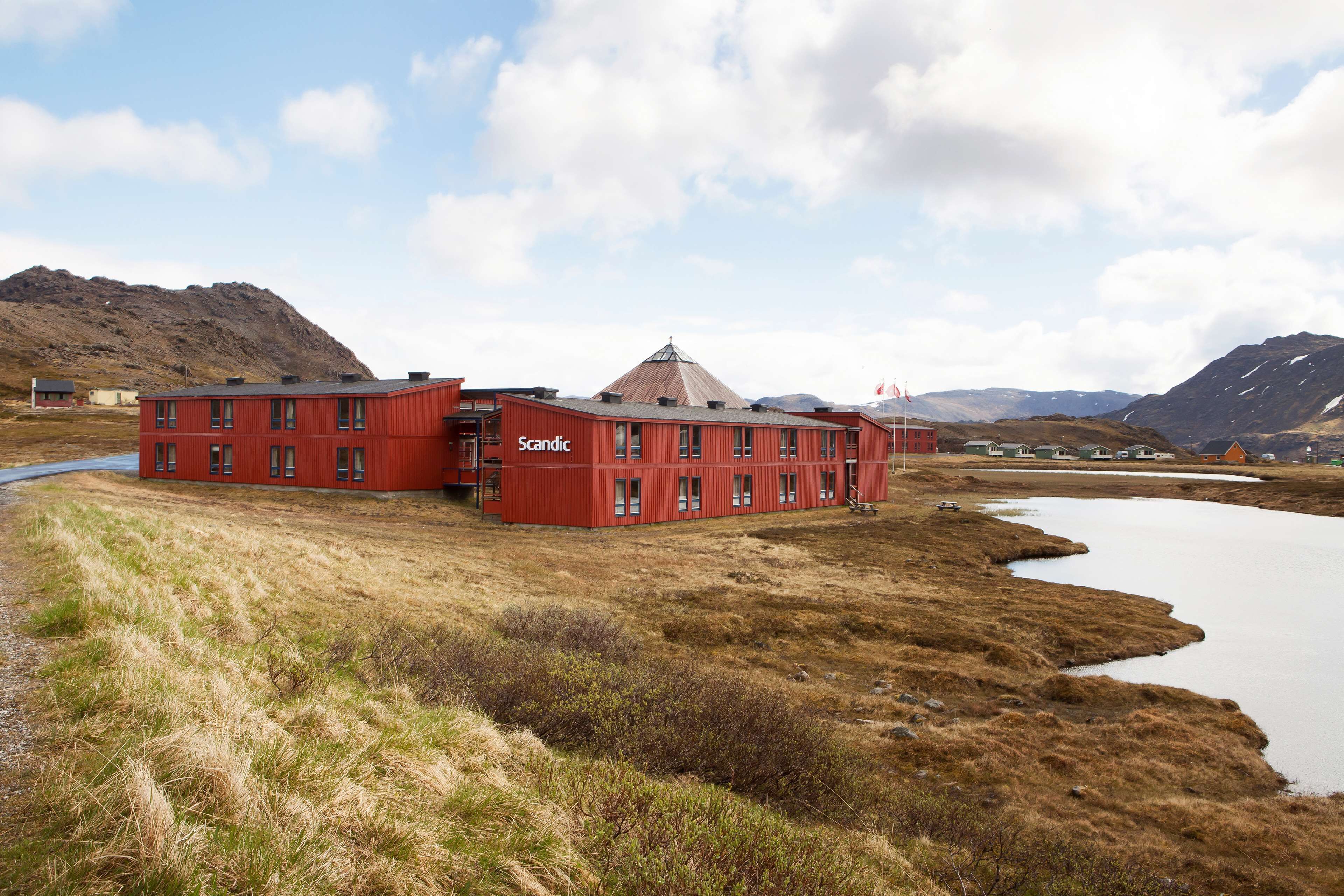 A red building named 'Scandic' with a unique triangular roof, situated by a calm lake with mountains in the background.