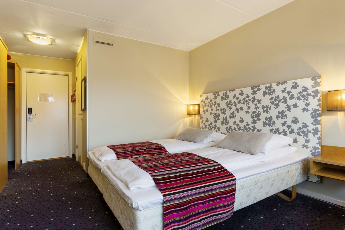 Habitación de hotel con cama, lámpara de noche, cabecera con estampado floral y suelo alfombrado.
