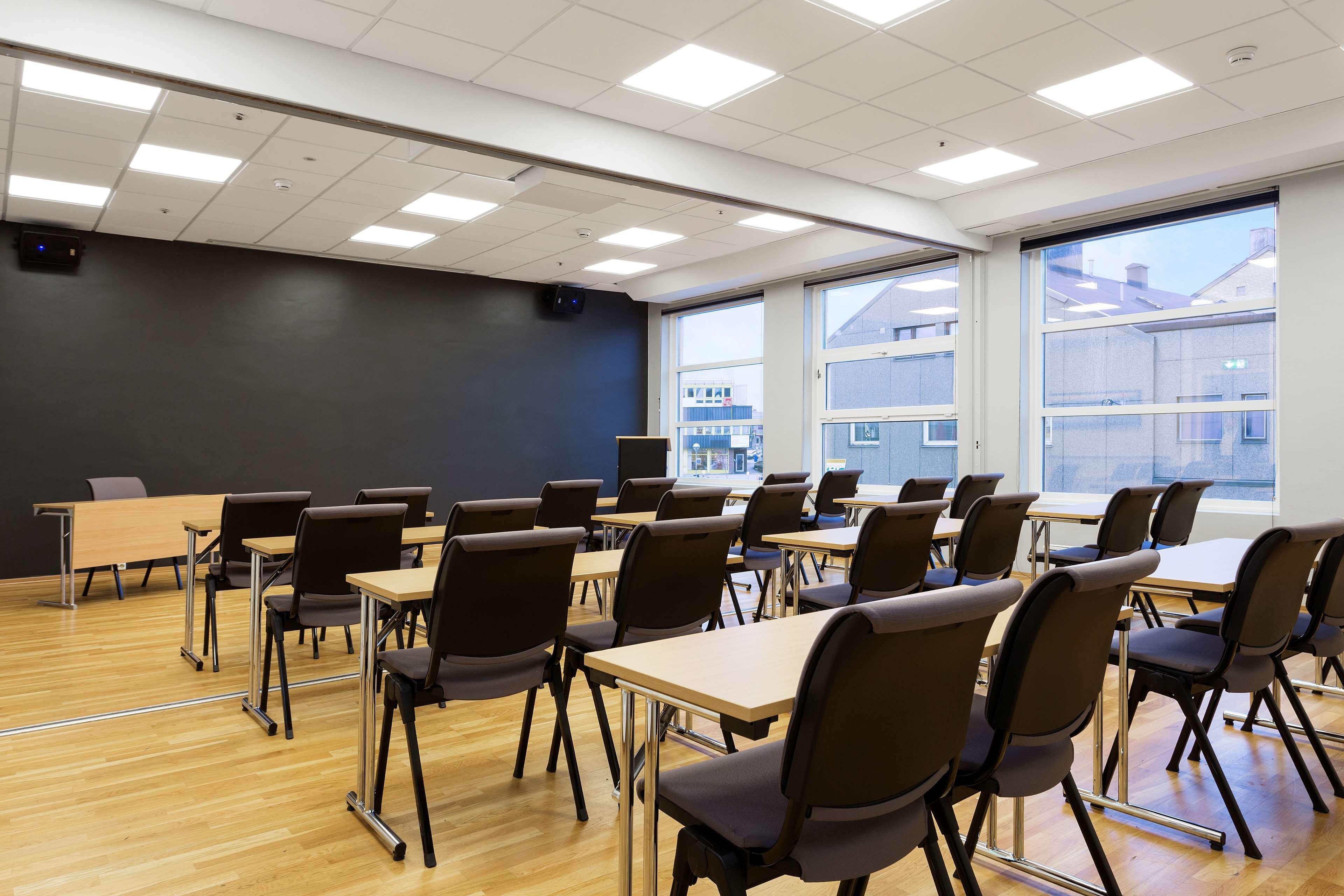 A classroom with rows of desks and chairs, large windows, and a projector on the ceiling.